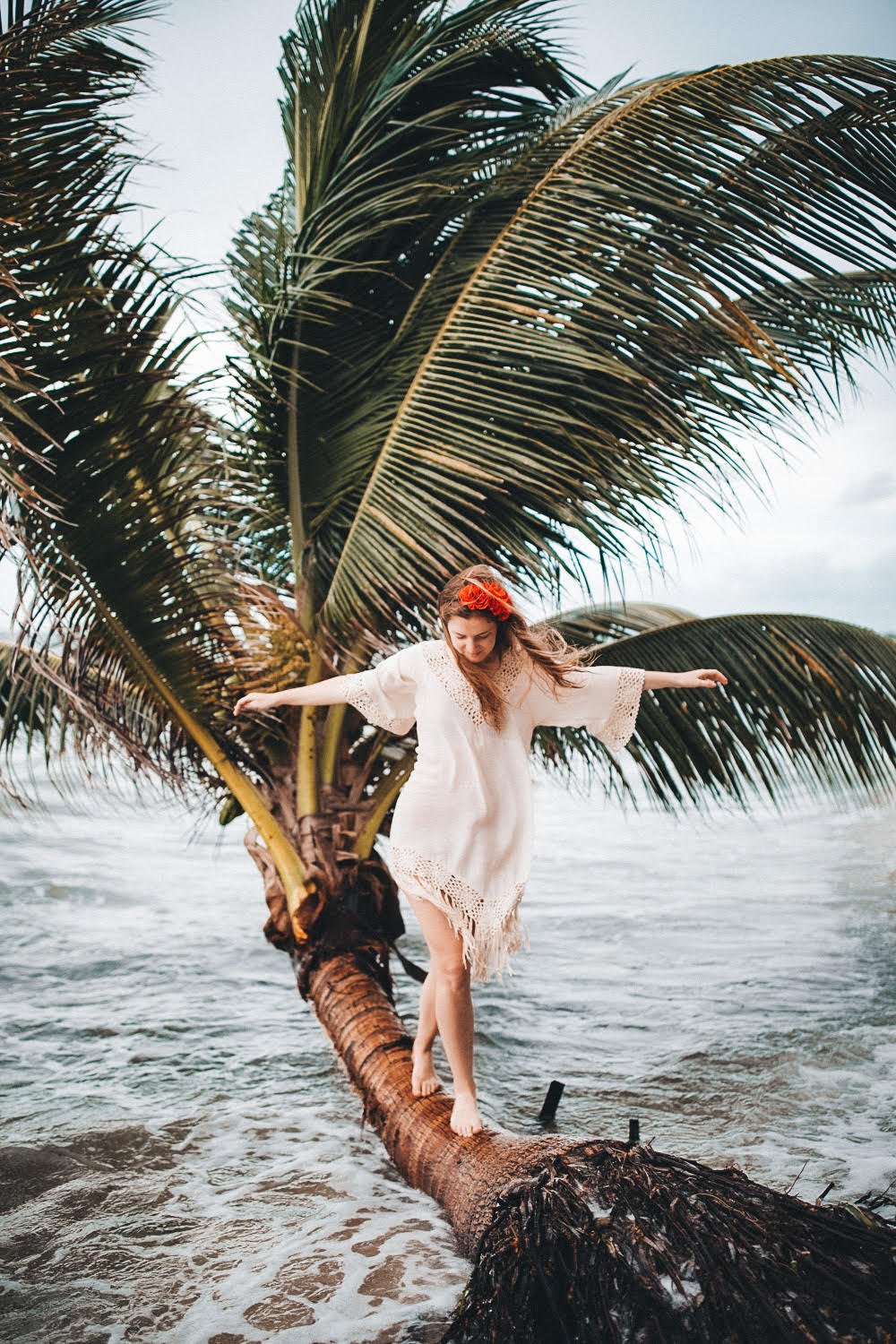 Woman in a white dress standing on a fallen palm tree in the ocean, with large palm tree fronds behind her and cloudy sky overhead.