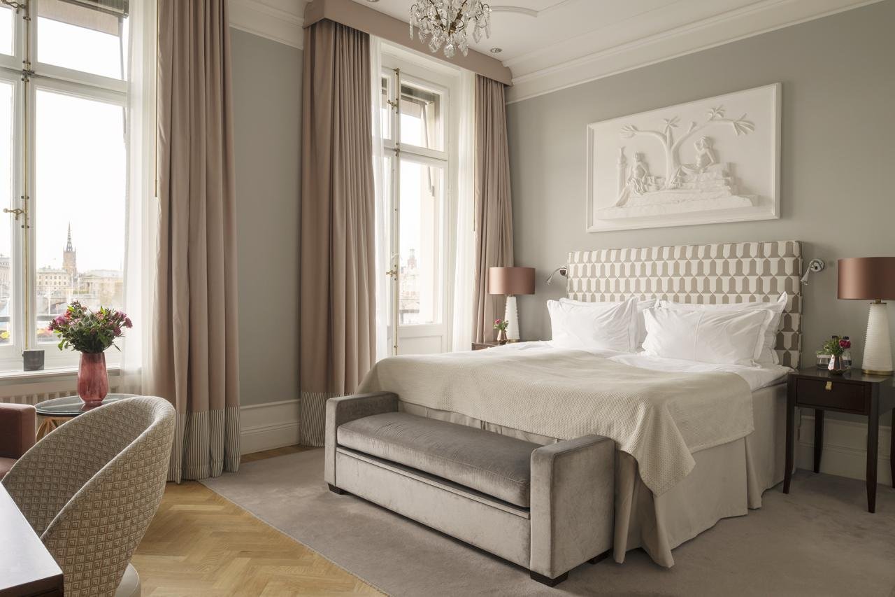 Elegant hotel room with large windows, beige curtains, a bed with a patterned headboard, white bedding, twin lamps on side tables, a bench at the foot of the bed, and city views outside.