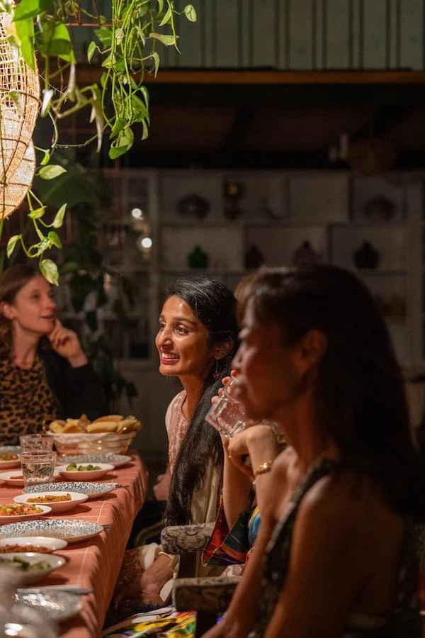 People sitting at a dining table with food, having a conversation, under hanging plants and warm lighting.