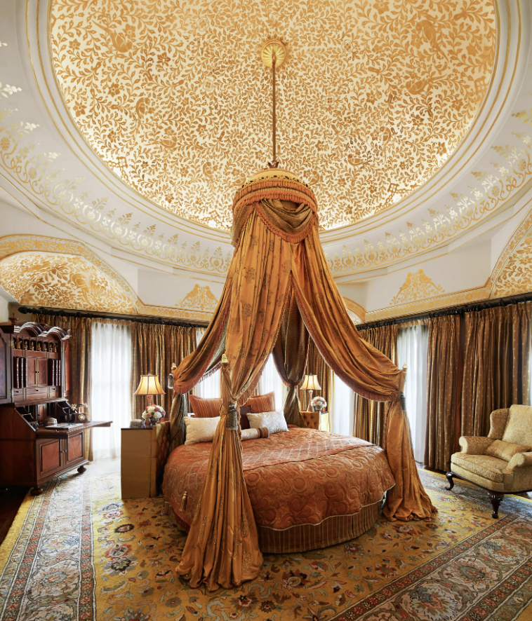 Luxurious bedroom with a round bed and gold canopy, ornate ceiling design, elegant armchair, and rich drapes.