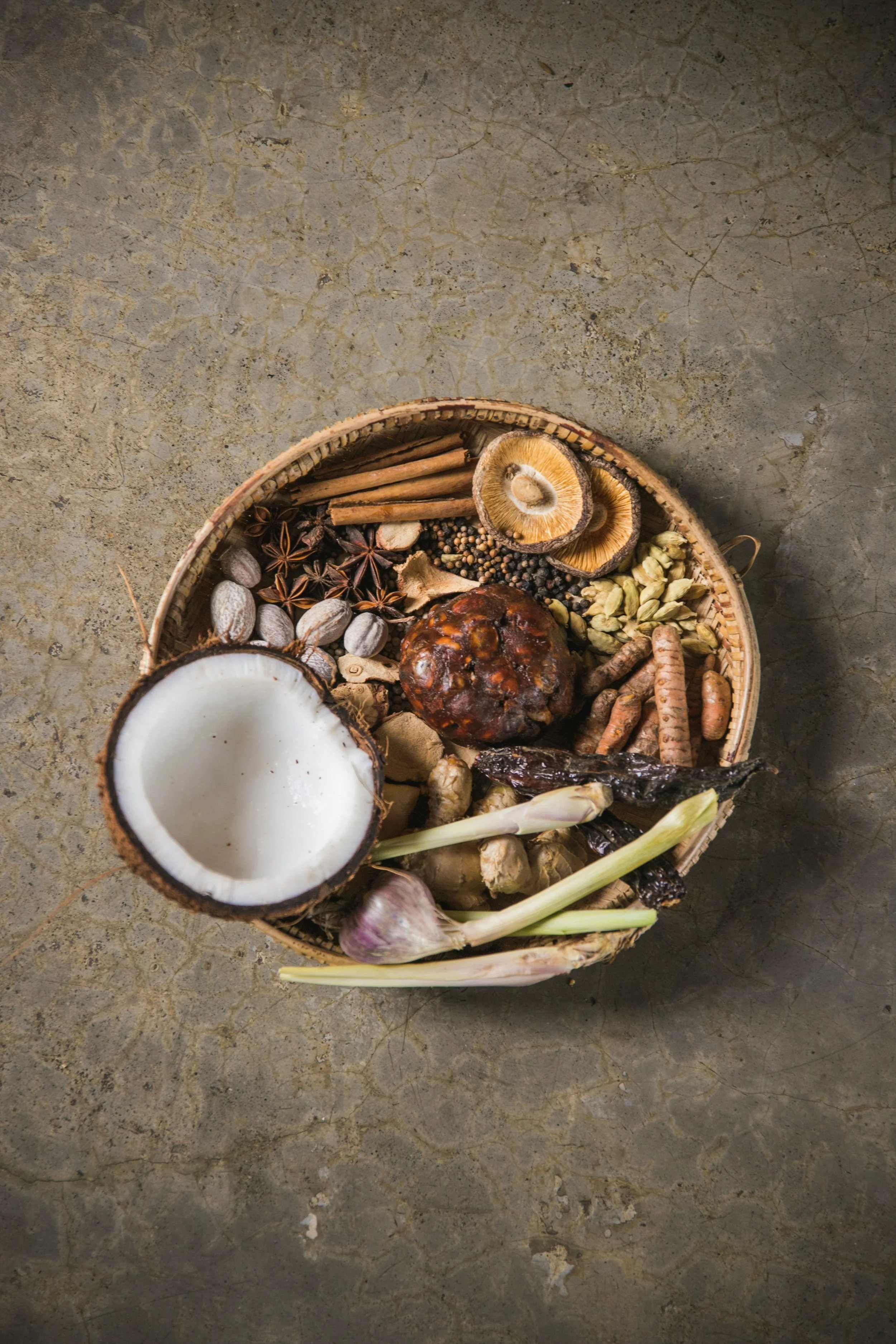 Assorted spices and herbs in a basket, including coconut, cinnamon sticks, star anise, lemongrass, garlic, ginger, dried peppers, dried mushrooms, cardamom pods, and black peppercorns, on a textured stone surface.