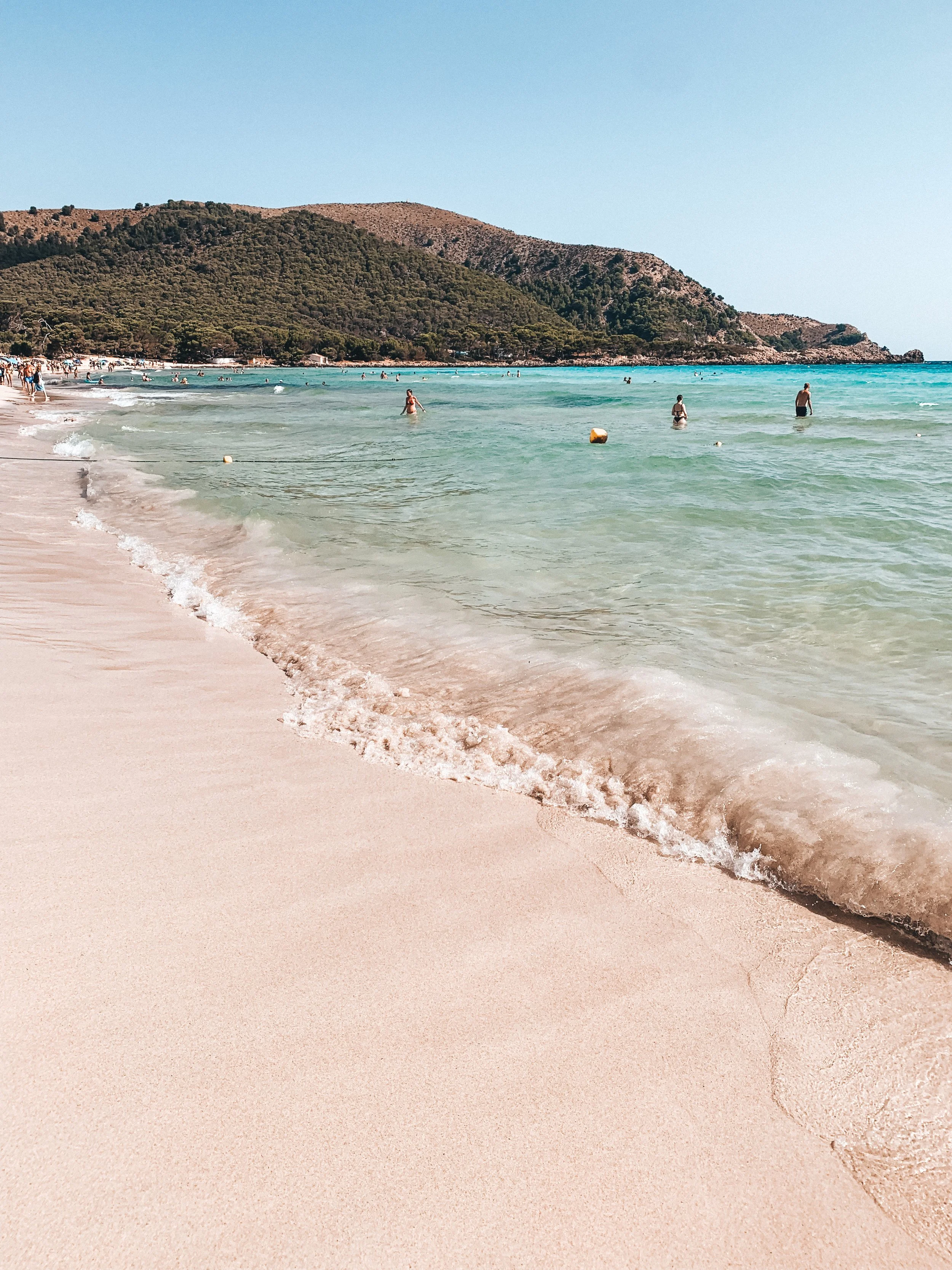 People swimming and relaxing at a sandy beach with clear turquoise water, with a hilly coastline in the background on a sunny day.