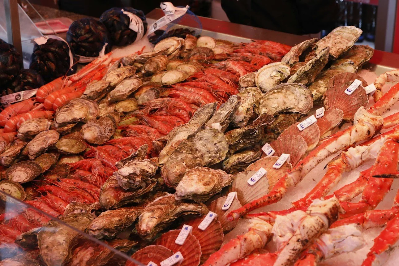 Display of various fresh seafood including oysters, scallops, prawns, scallop shells, and crab legs on a market counter.