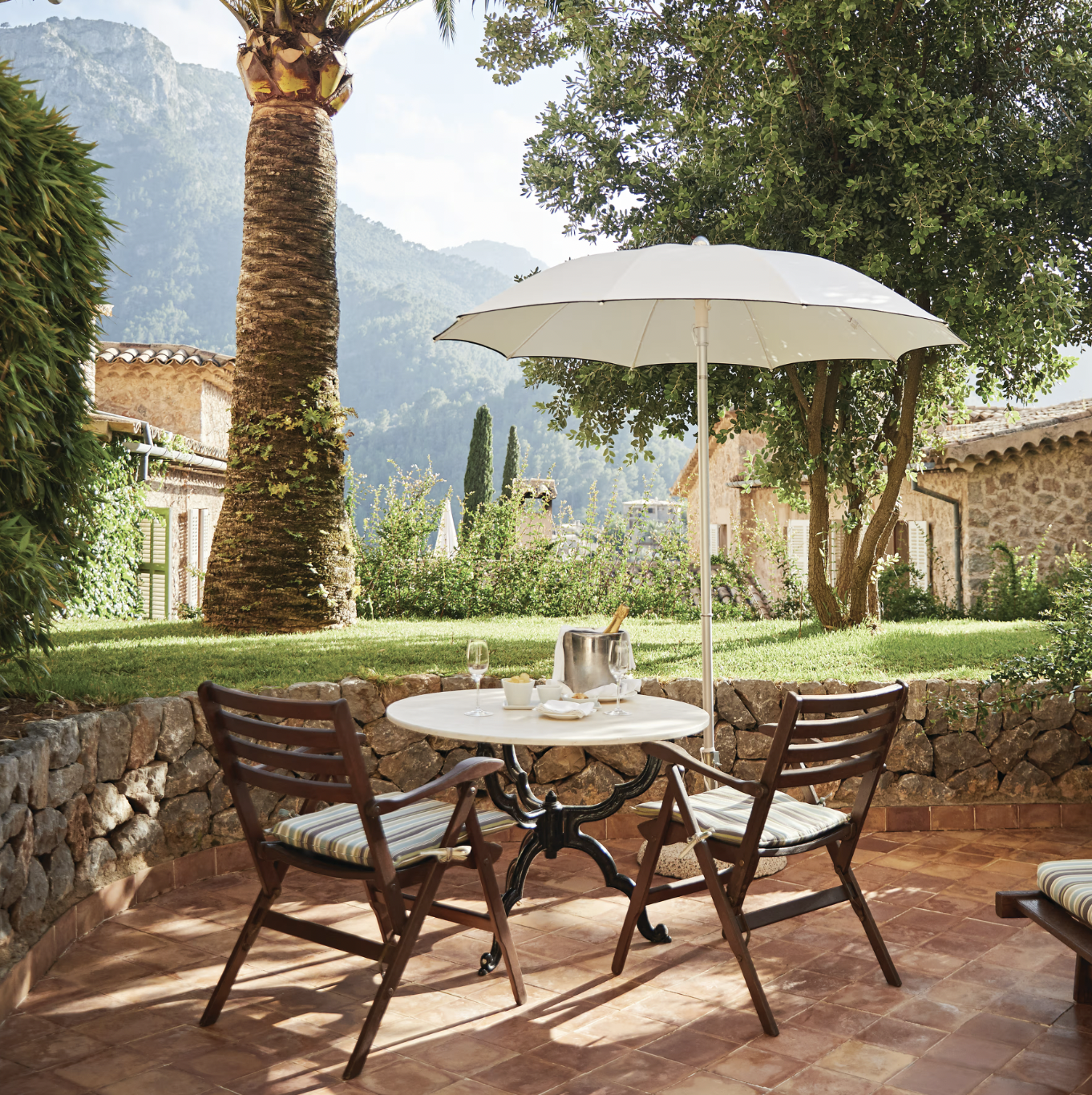 Outdoor patio with a table and two chairs, an umbrella providing shade, and scenic mountains and greenery in the background.