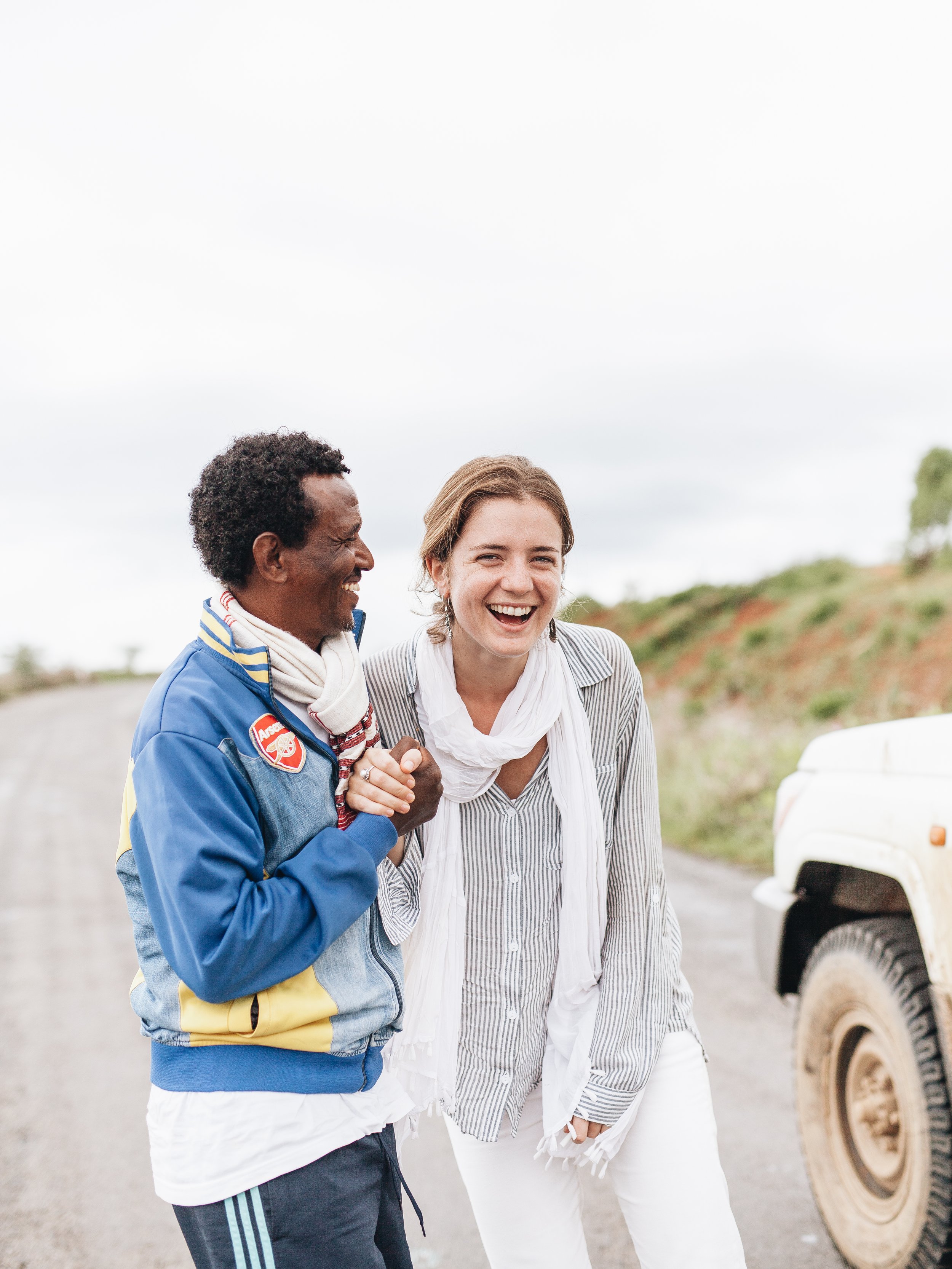 Two women sharing a joyful moment outdoors, one wearing an Arsenal jacket and the other in a striped shirt and white scarf, standing near a white vehicle with a rural background.