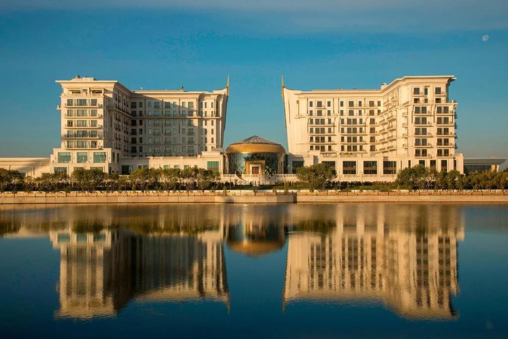 Two large modern hotels with unique architectural designs by a body of water, with their reflections visible in the water, under a clear blue sky.