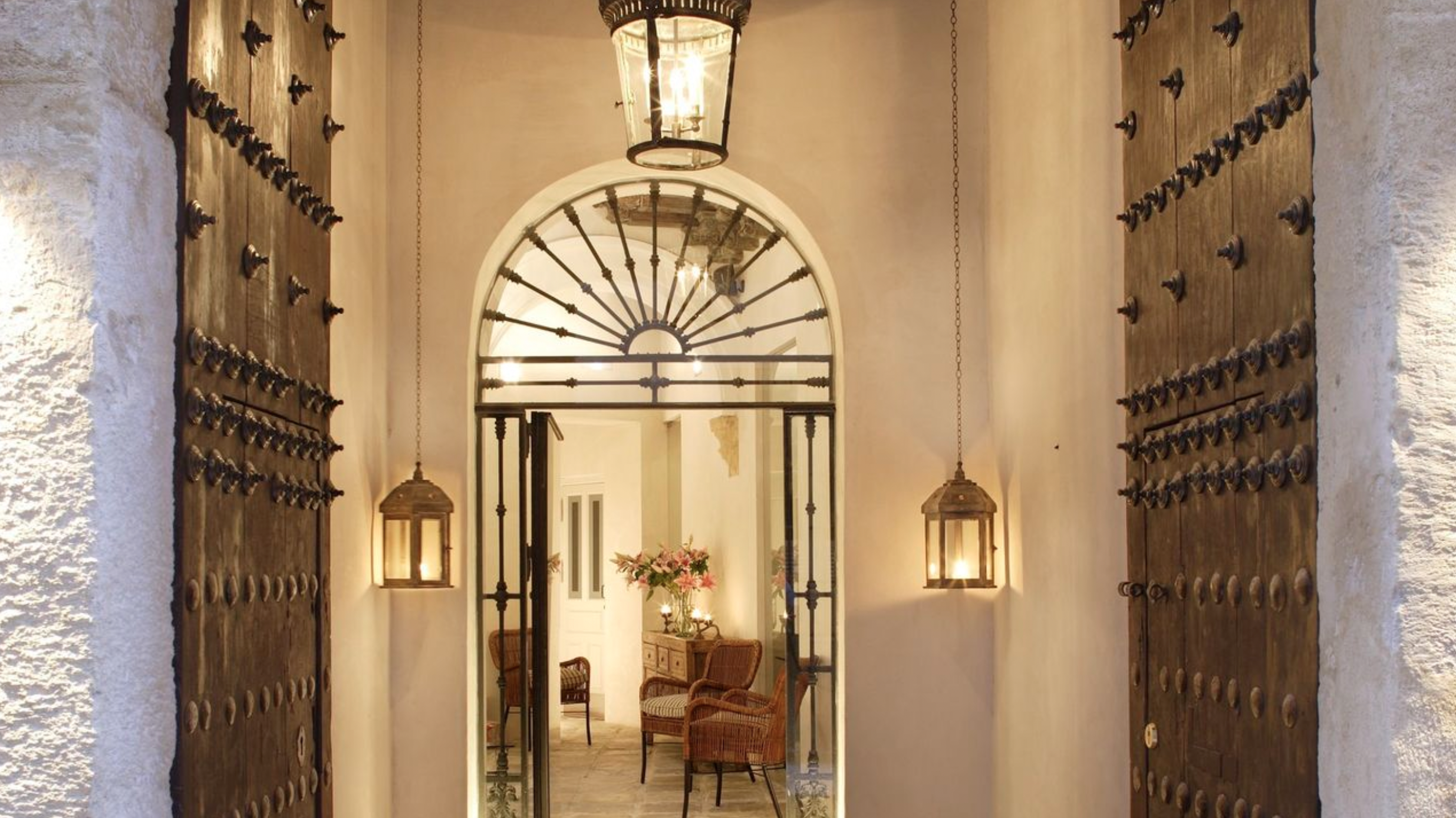 Elegant entrance hallway with large wooden doors, lantern lights, and arched gateway leading to a cozy seating area with wicker chairs and flowers.