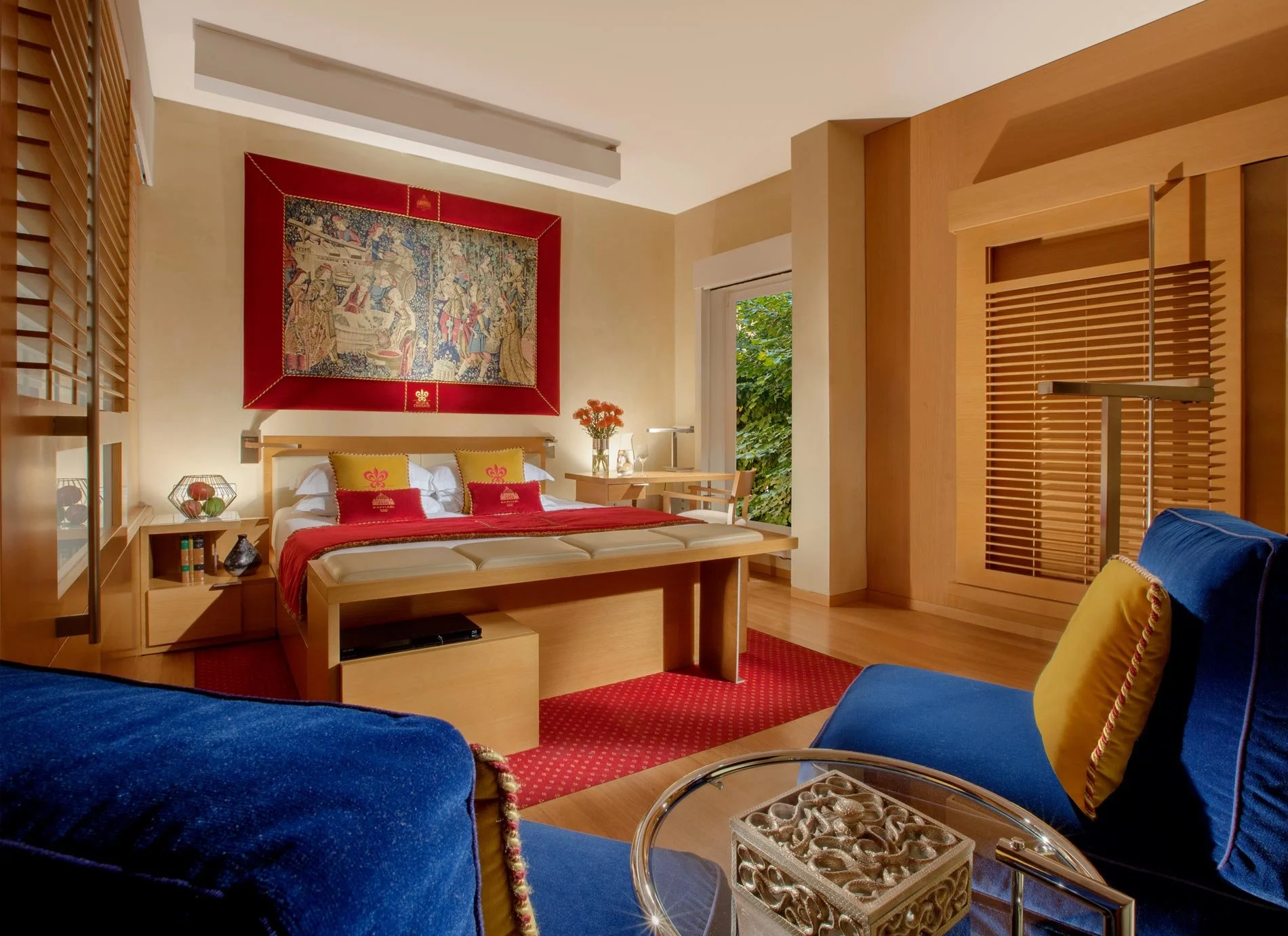 Elegant hotel room with a large bed, blue chair, decorative tapestry, and wooden furniture.