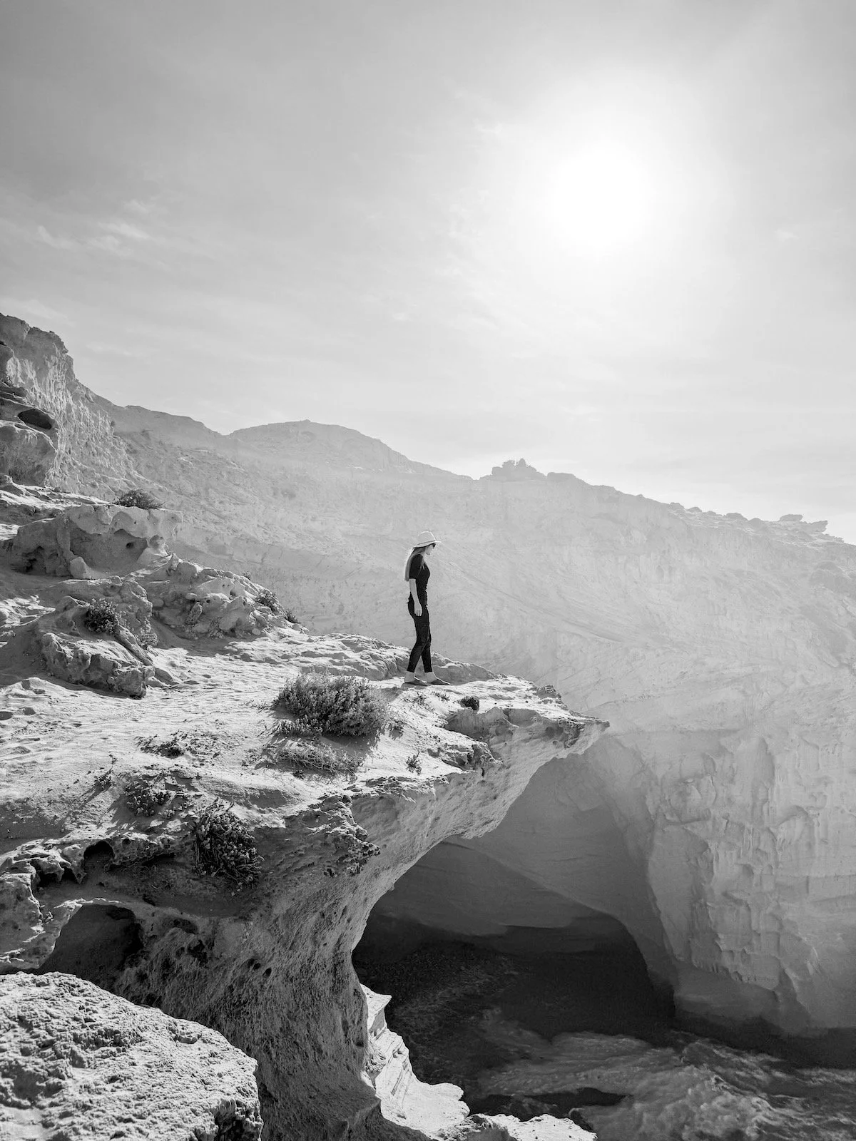 A person standing on a rocky outcrop overlooking a cave or abyss in a dry, mountainous landscape with the sun shining in the sky.