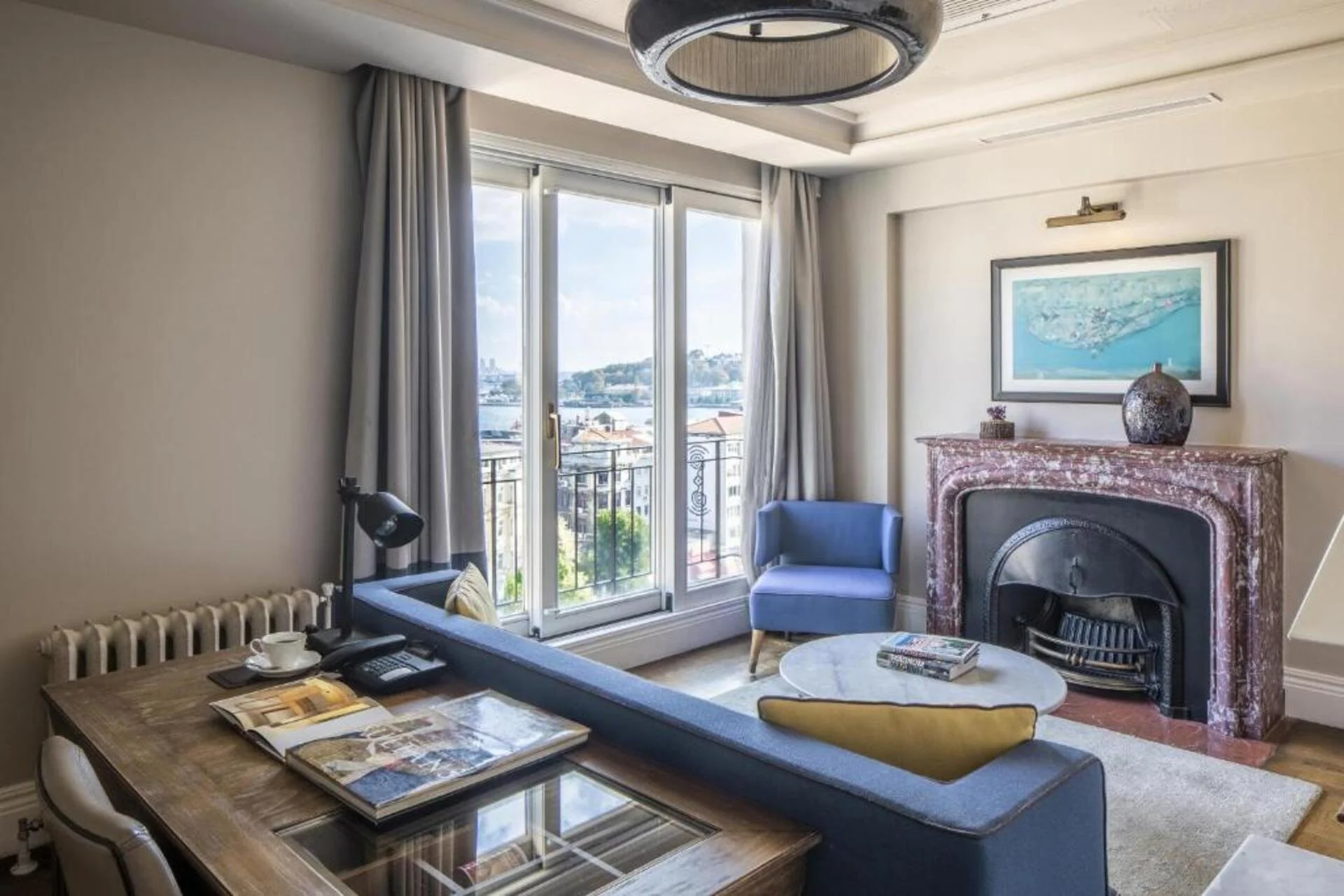 A cozy living room with a large window overlooking a view of city buildings and water. The room features a blue armchair, a marble coffee table with magazines, a fireplace with decorative items on top, and a wooden desk with books and a cup of coffee.