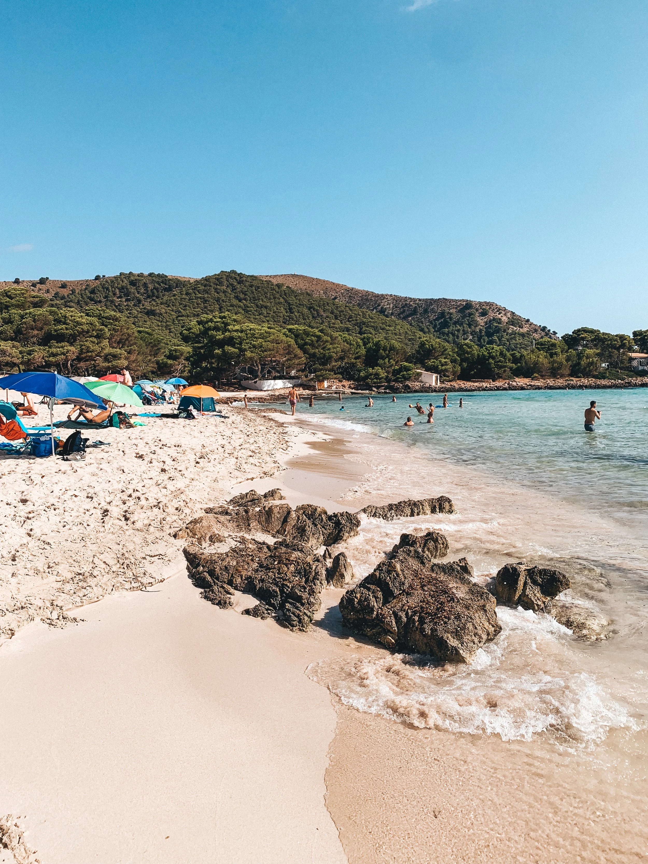 A sunny beach with sandy shoreline, rocks in the water, beachgoers swimming and relaxing under umbrellas, with trees and hills in the background under a clear blue sky.