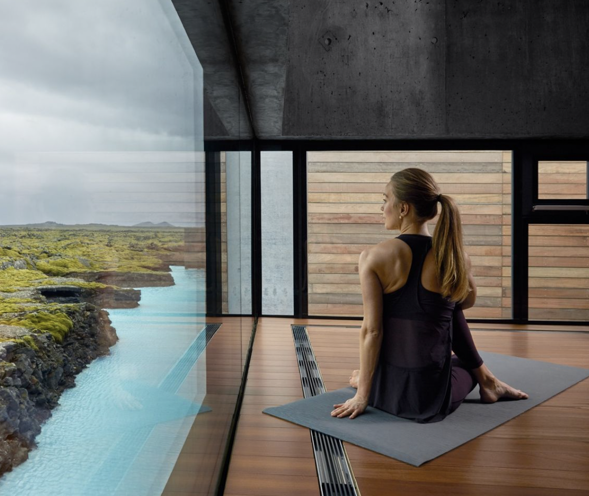 Woman practicing yoga indoors on a mat, overlooking a scenic view of water and rocks through large windows.