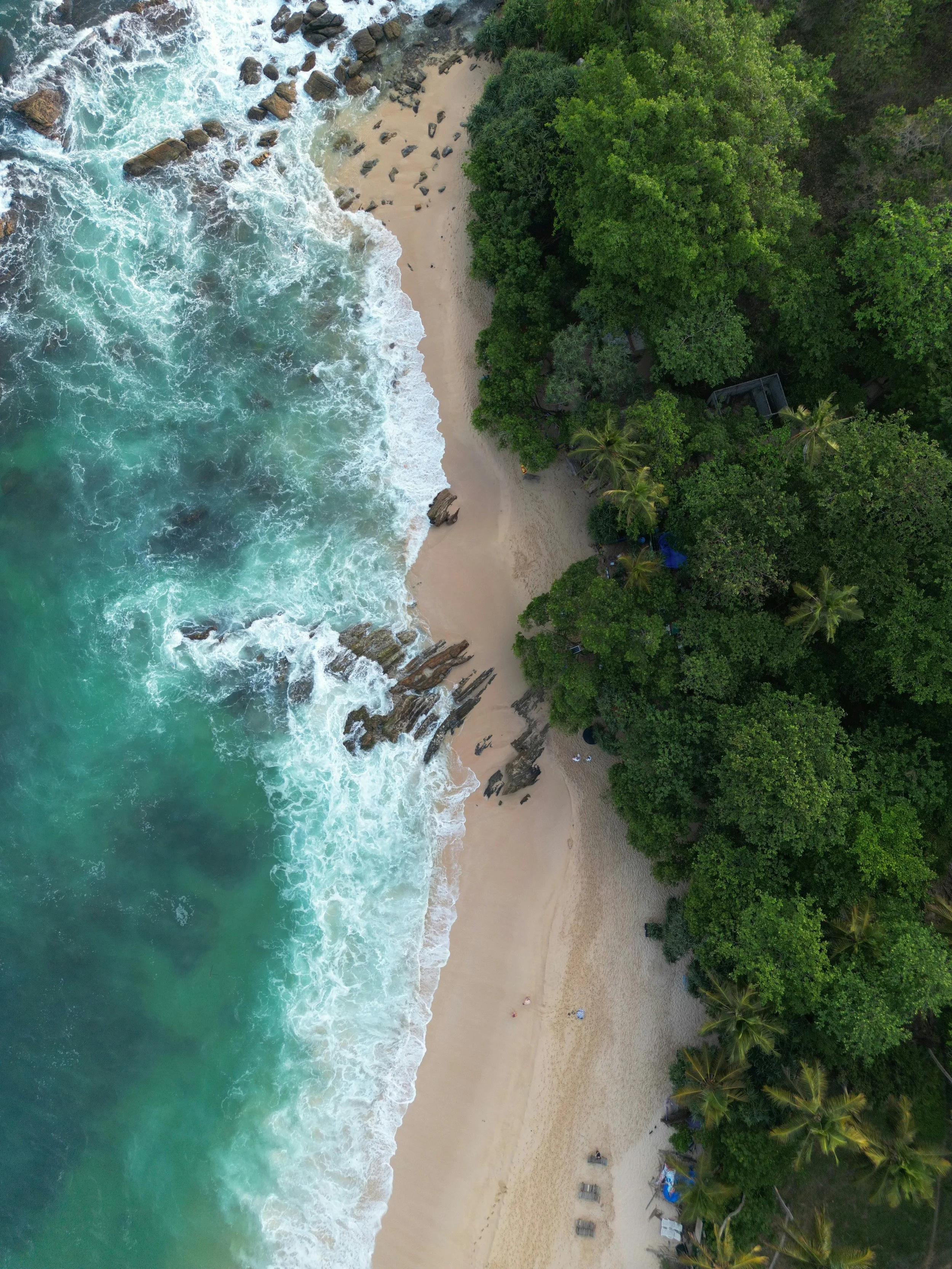 Aerial view of a sandy beach with turquoise waves crashing against the shore, bordered by lush green forest.
