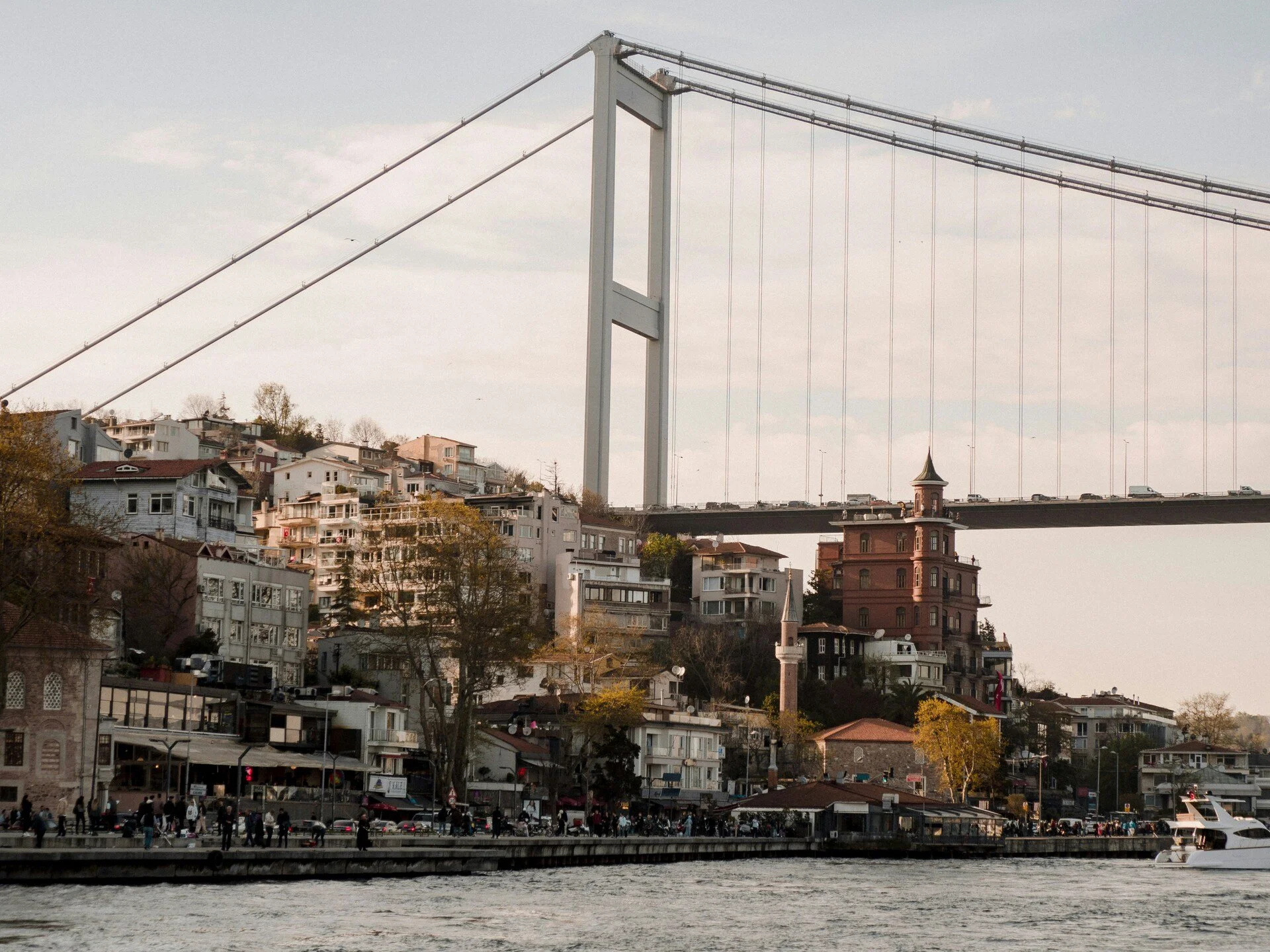 View of a city hillside with numerous residential buildings, a large bridge crossing over, and a waterfront with people and boats.