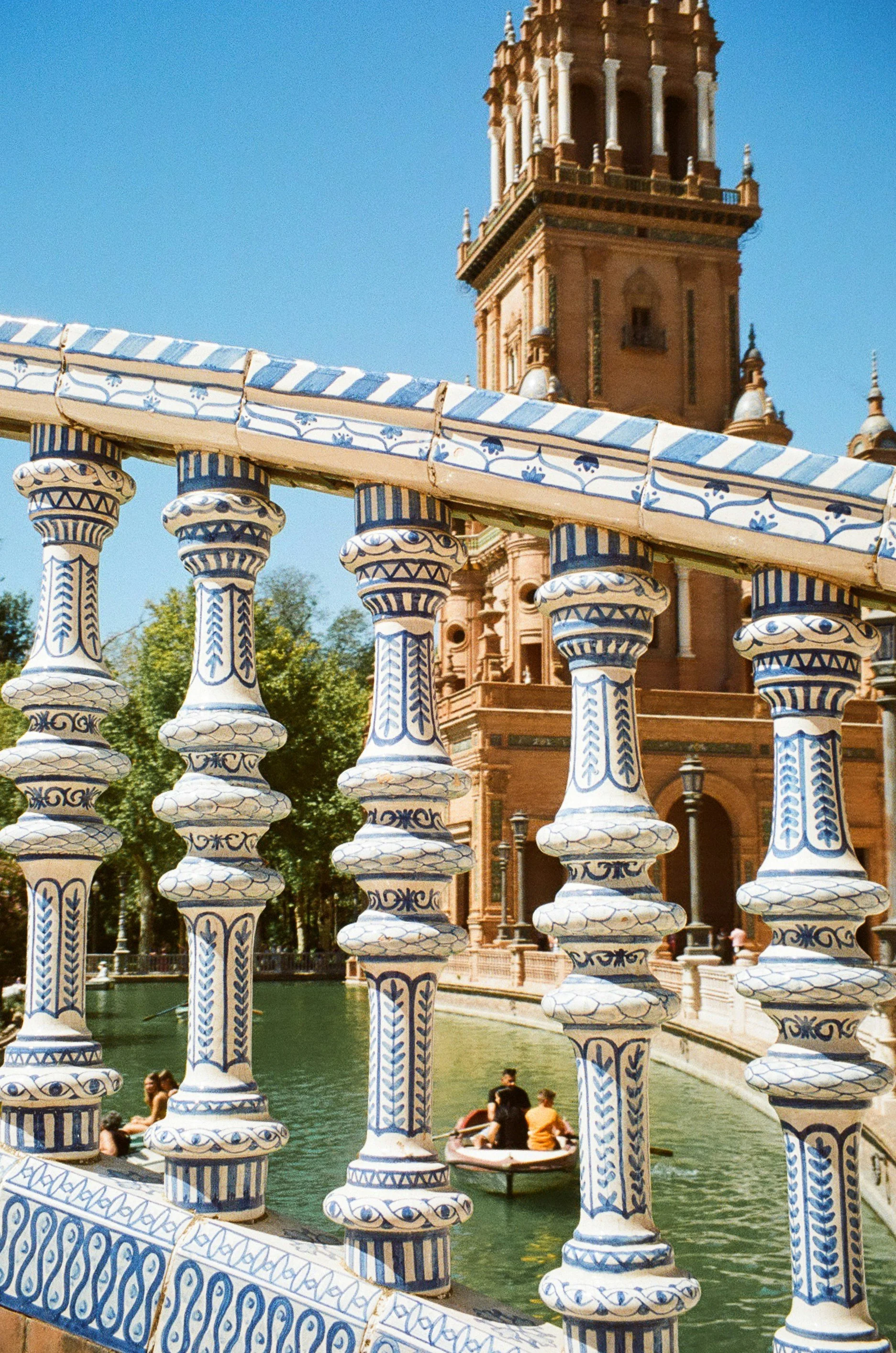 Ornate balustrade with blue and white ceramic design in Plaza de España, Seville, featuring a water canal and a tower.