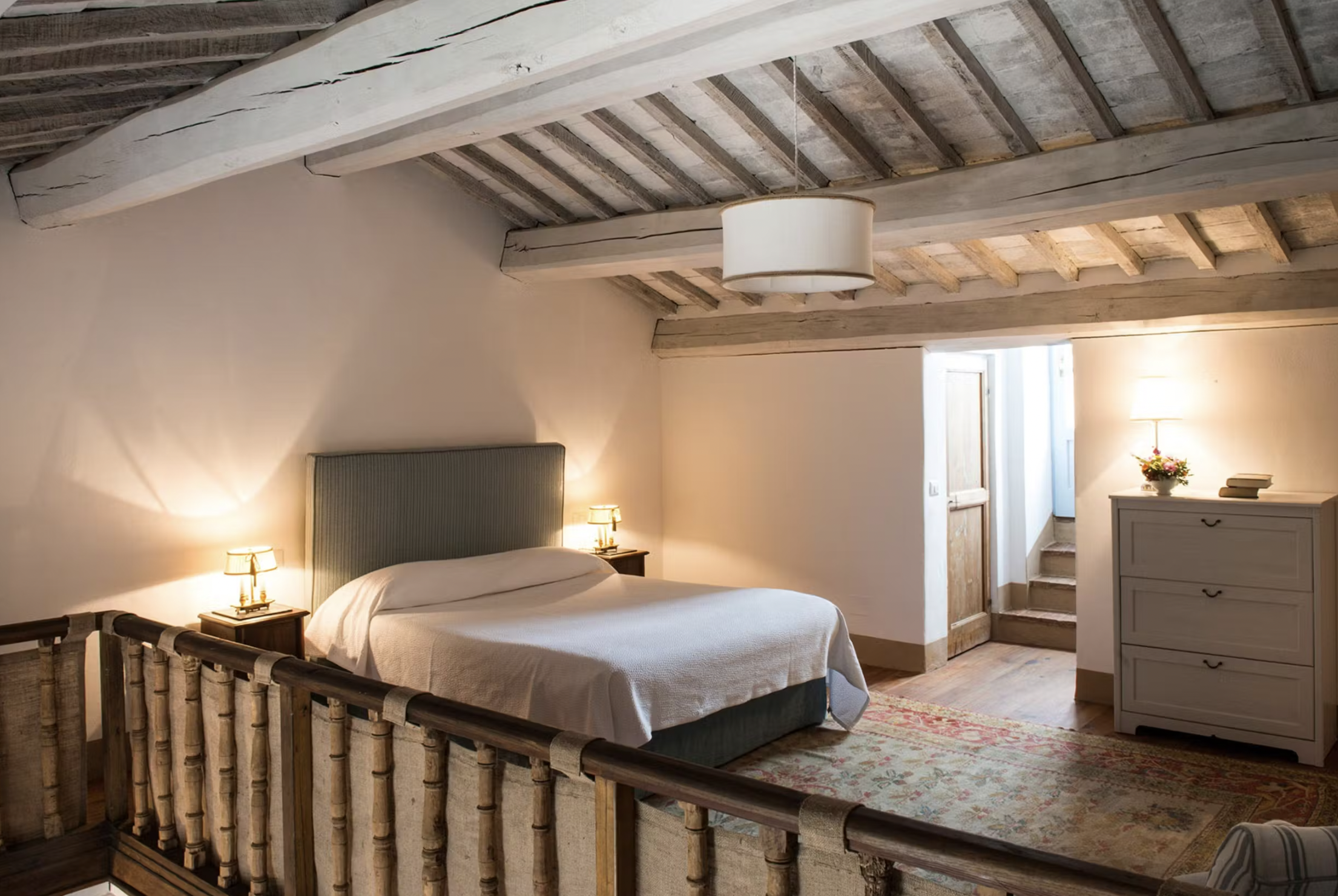 Cozy attic bedroom with wooden beams, a double bed, nightstands, soft lighting, a dresser, and a rug.