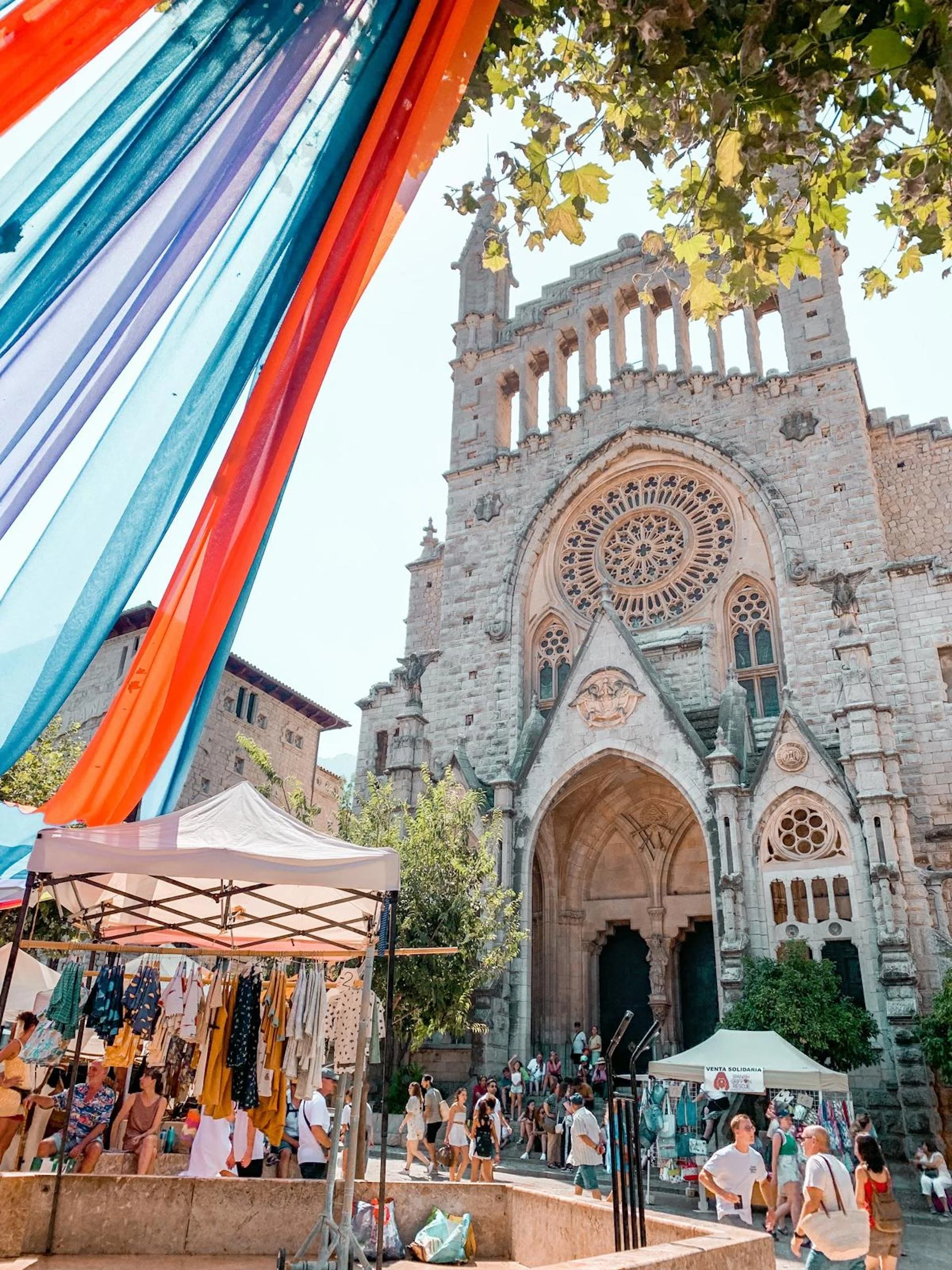 Crowd of people in front of a historic stone church with intricate gothic architecture, surrounded by market stalls and colorful banners.