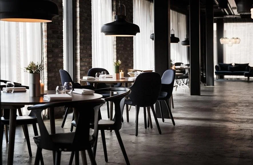 Interior of a modern restaurant with round tables, black chairs, large pendant lights, and floor-to-ceiling windows with sheer curtains.