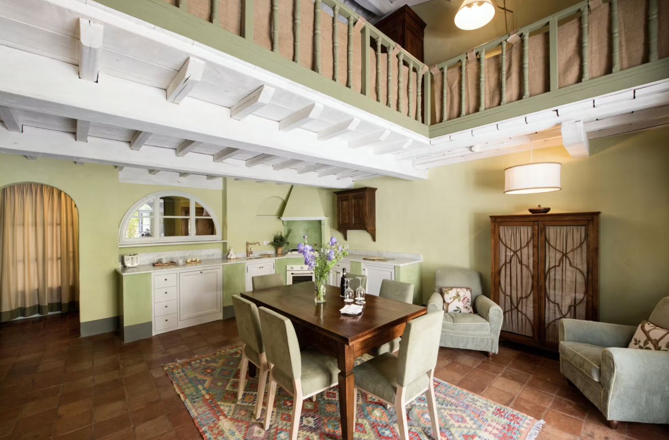 Cozy kitchen and dining area with vintage decor, featuring a wooden dining table with four upholstered chairs, armchairs, a colorful rug, and an overhead loft with a railing.