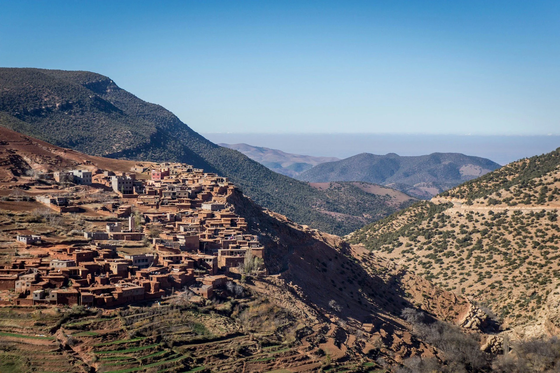 A mountain village built on a hillside with terraced fields and rugged mountains in the background under a clear blue sky.