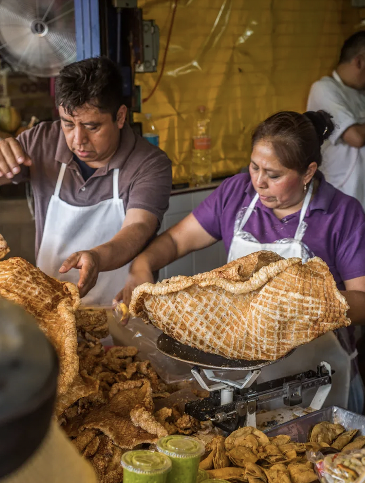 Two people weighing a large piece of chicharrón at a food stall, with jars of green sauce in the foreground.