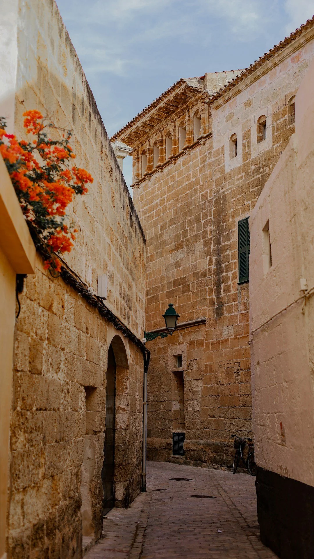 A narrow cobblestone street lined with old stone buildings, including a tower with medieval architectural features, a green street lamp, a bicycle, and vibrant orange flowers against a partly cloudy sky.