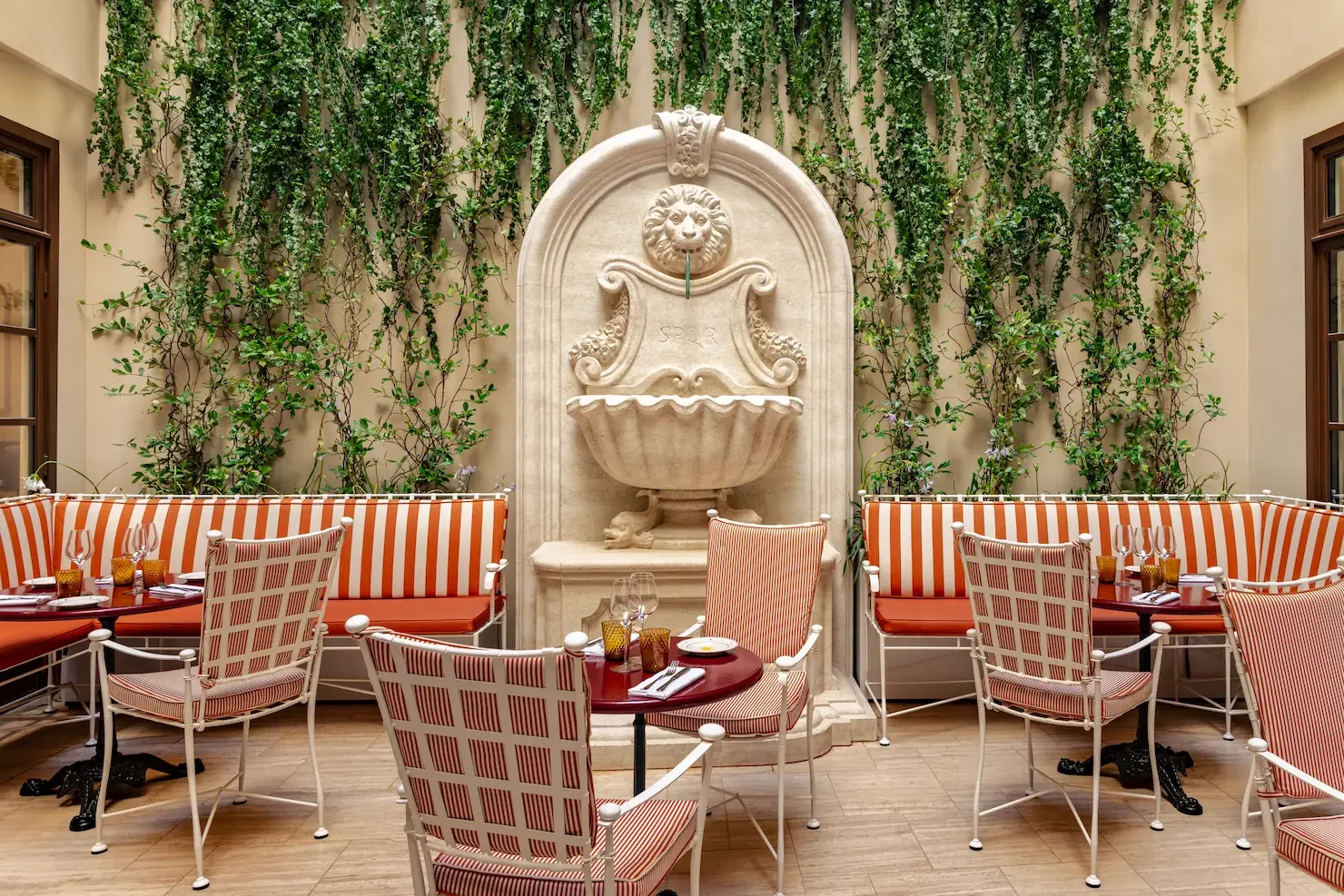 A waiter standing on a terrace with pink flowers and tables, holding a tray with glasses. There's a building with classical architecture in the background.