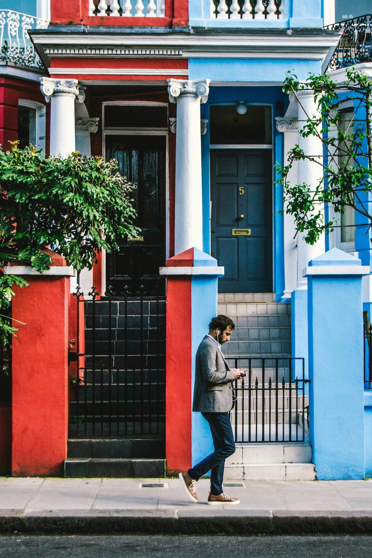 A man with dark hair, wearing a gray blazer, dark pants, and sneakers, stands on a city sidewalk, looking at his phone. Behind him are colorful houses with red, blue, and white facades, white columns, and stairs leading to their doors.