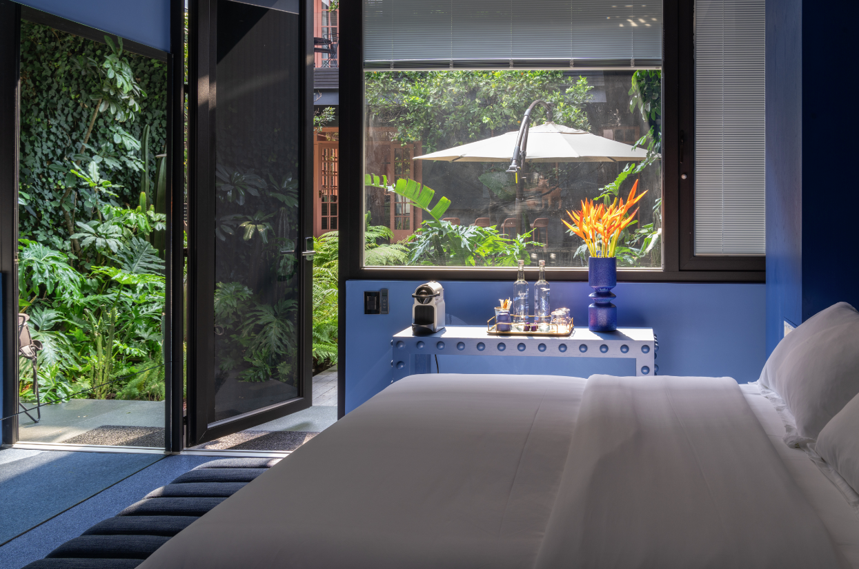 A modern bedroom with blue walls features an open glass door leading to a lush green garden. Inside, a neatly made white bed is visible beside a small table holding a coffee machine, water bottles, and candles. Bright orange flowers in a blue vase add a touch of color.