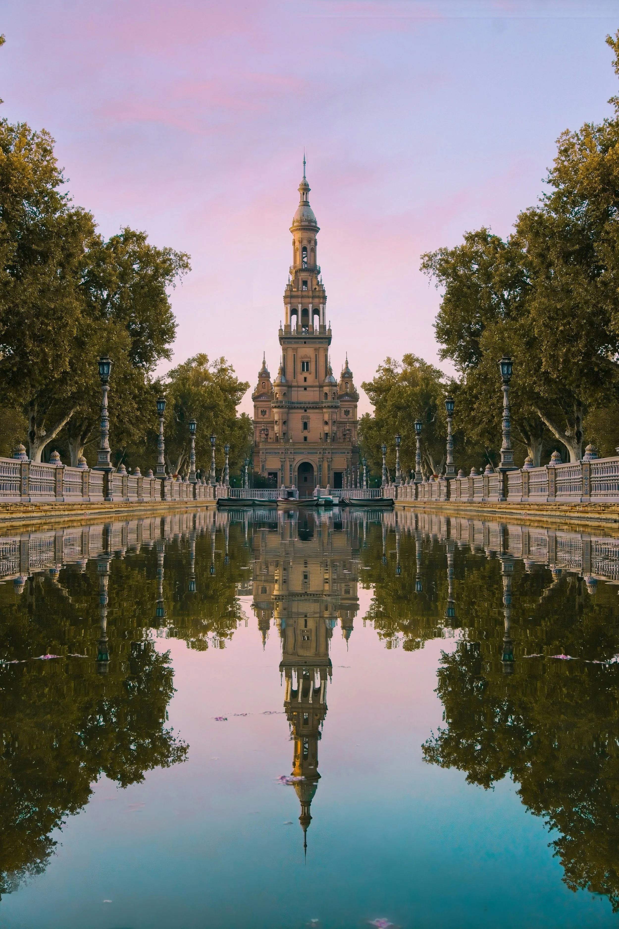 A tall historic building with a tower, reflected in a pond, surrounded by trees and lampposts, under a pink sky.