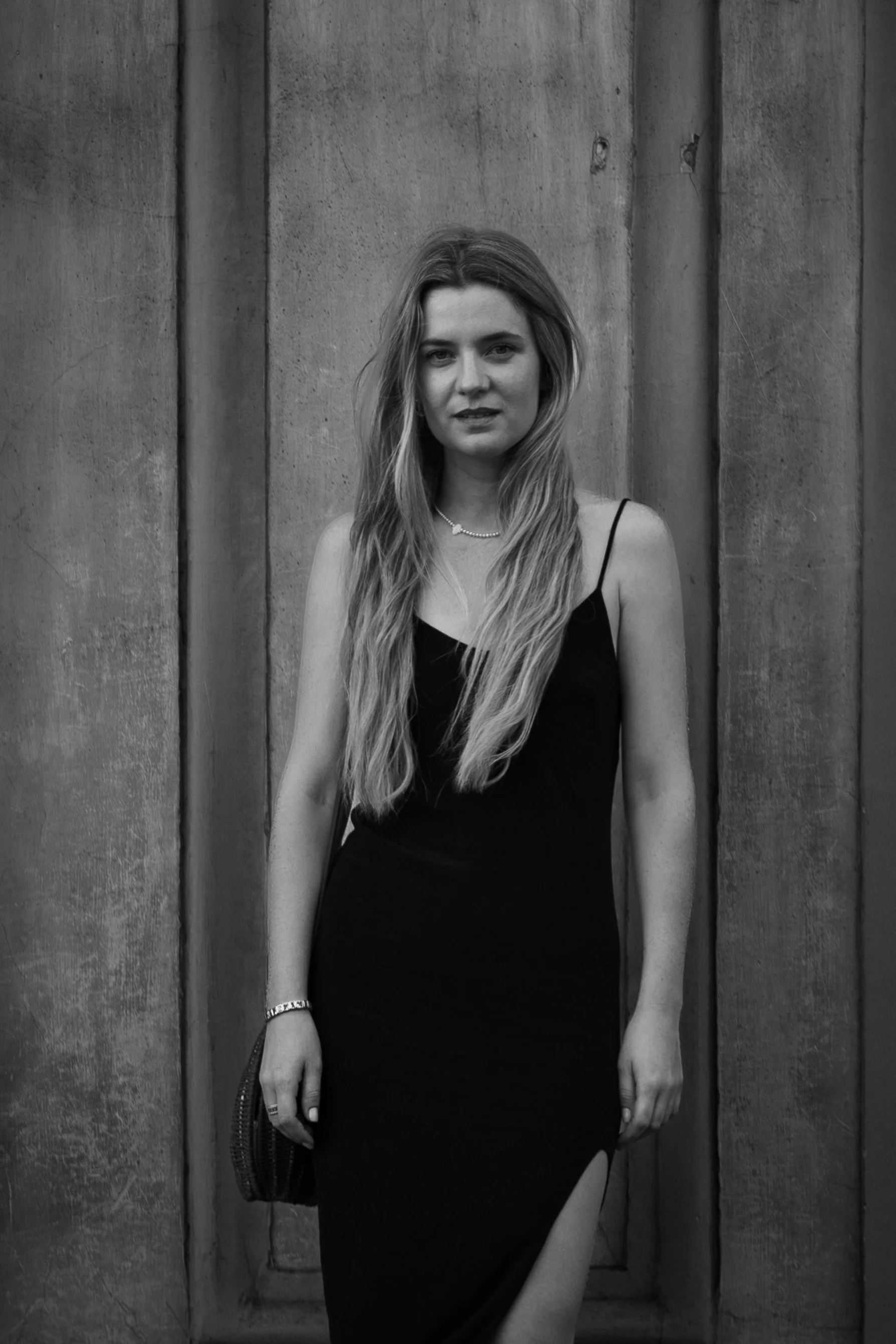 A young woman with long hair wearing a black dress with thin straps and a slit, standing in front of a textured wall.