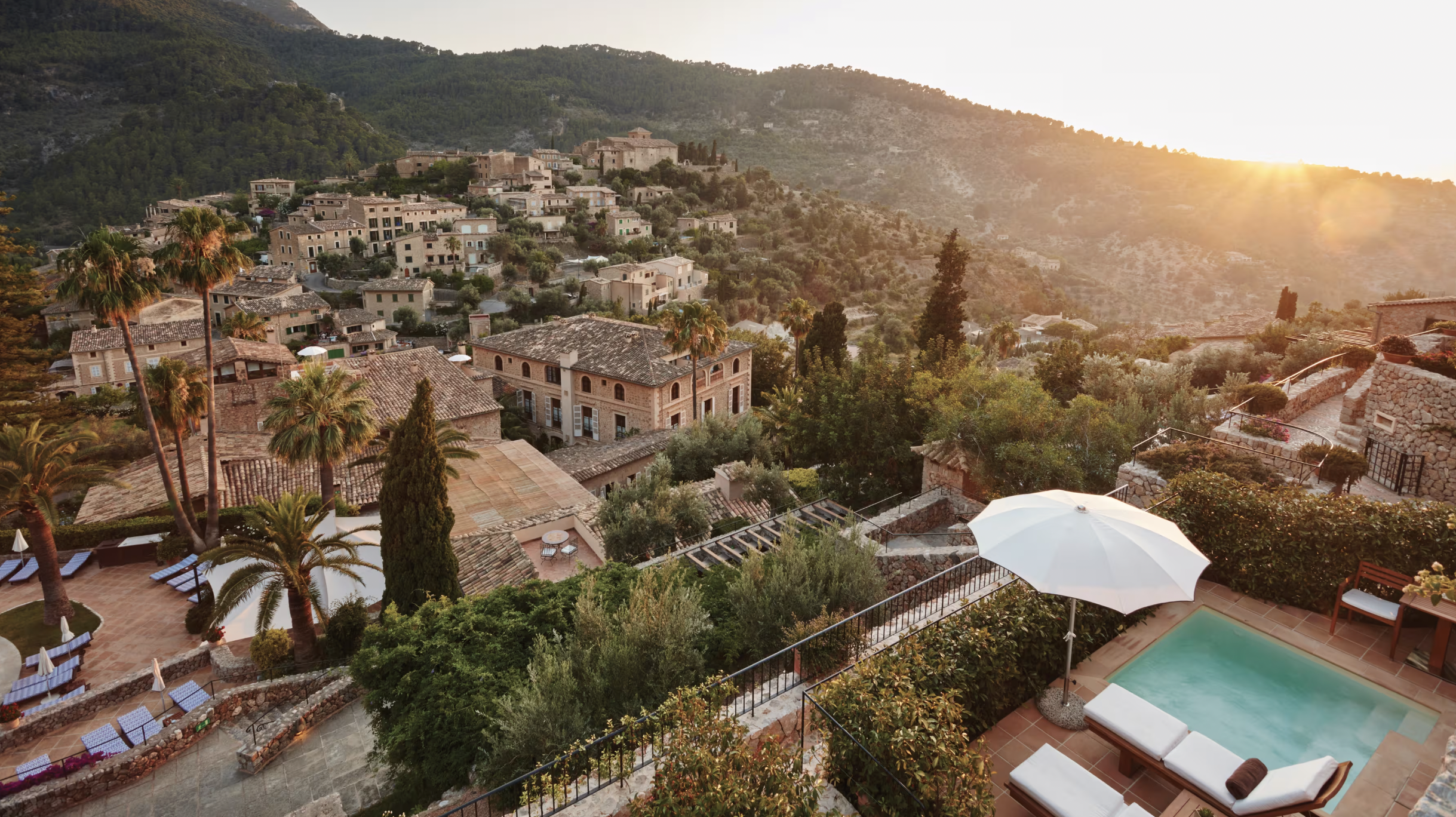 A hillside village at sunset with terracotta-roofed buildings, palm trees, lush greenery, and a patio with a small pool and white umbrellas in the foreground.