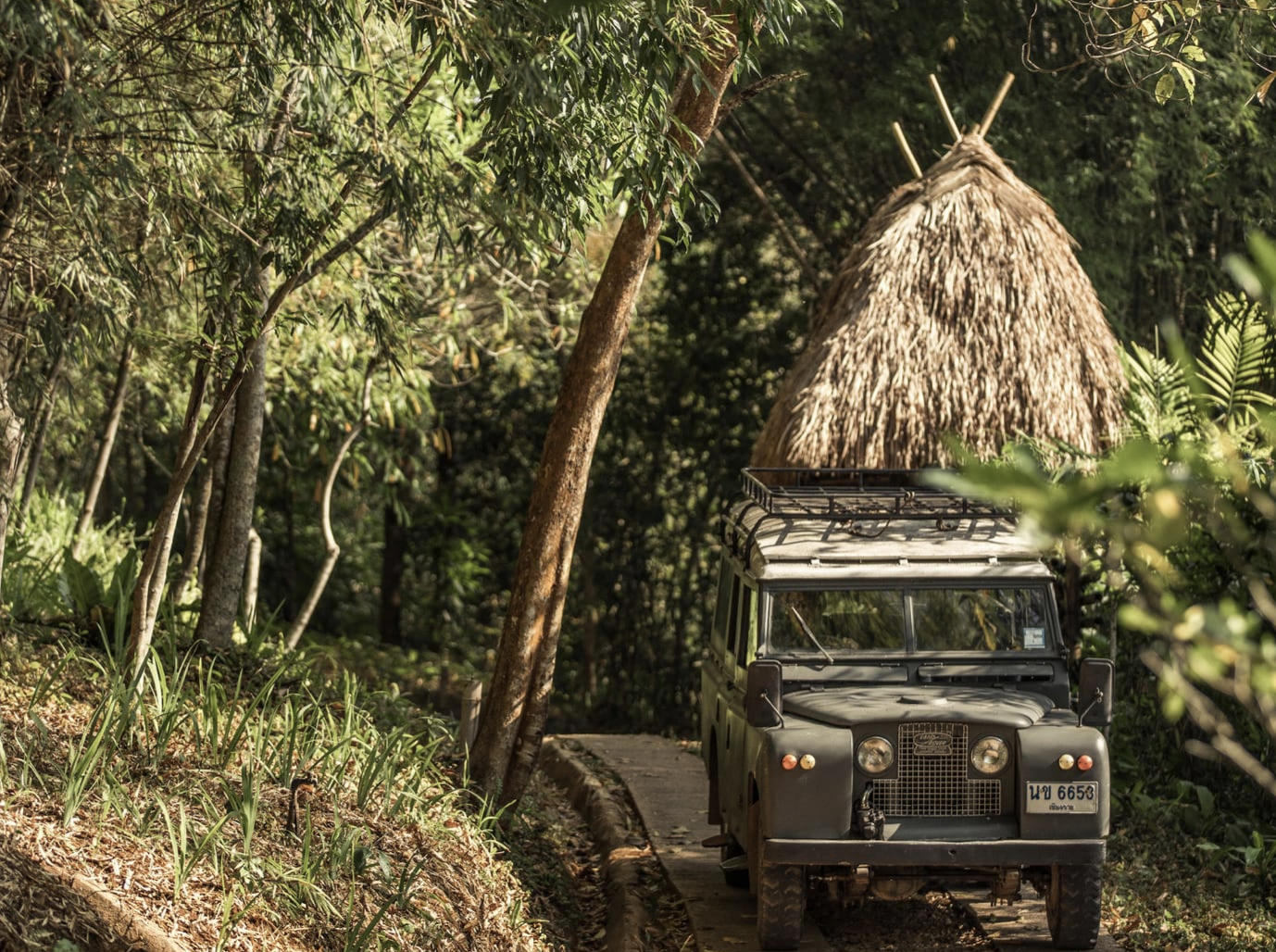 Vintage Land Rover on a narrow forest path next to a straw hut, surrounded by dense greenery.