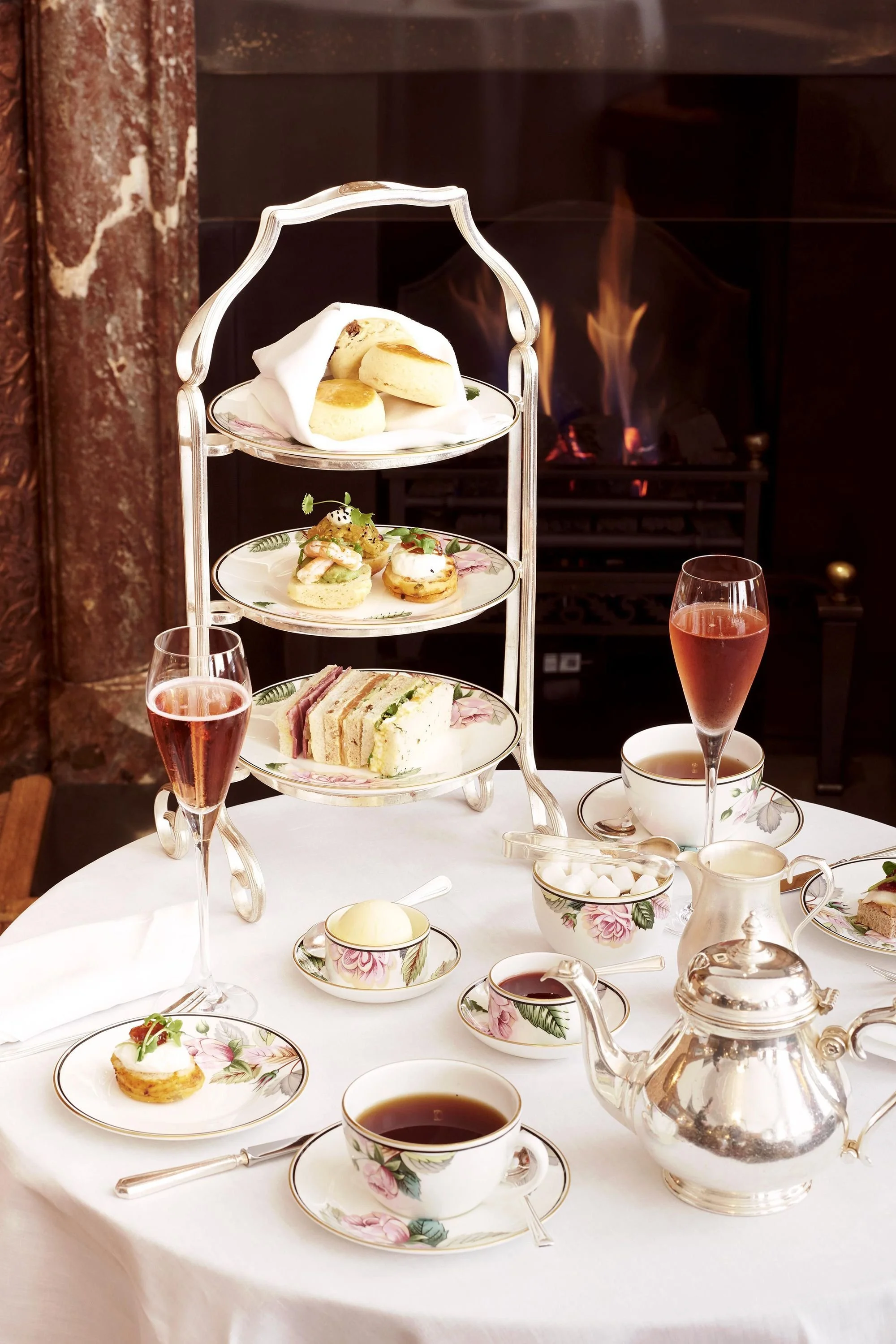 Elegant afternoon tea with assorted finger sandwiches, scones, pastries, and tea served in fine china with a silver teapot, set on a white tablecloth with a fireplace in the background.