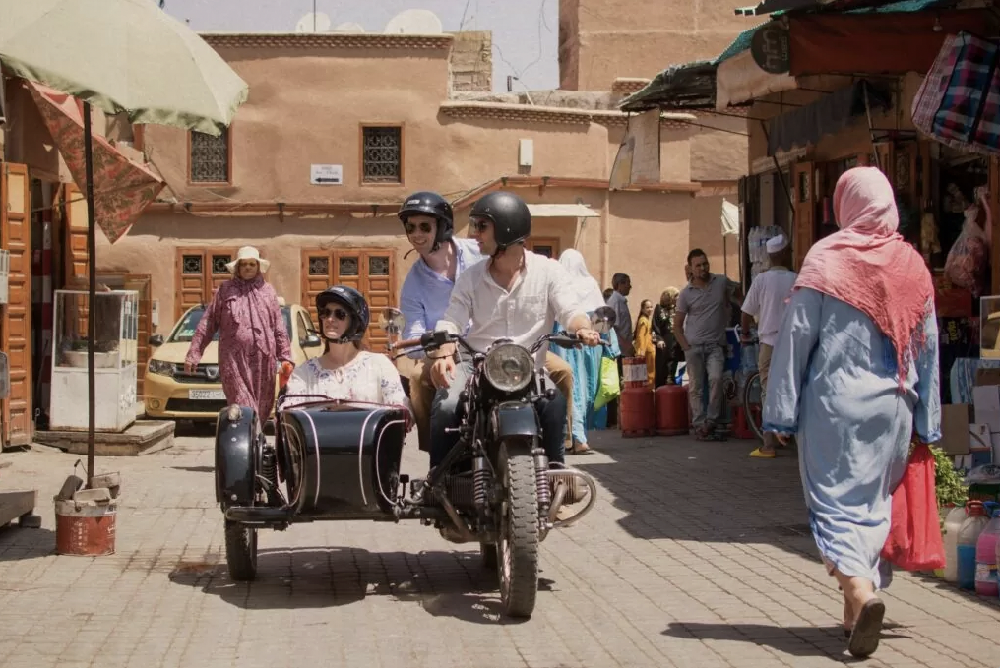 People riding a motorcycle with a sidecar through a busy marketplace. The driver and passenger wear helmets, and the sidecar passenger is a woman with sunglasses. Market stalls and pedestrians are visible in the background.
