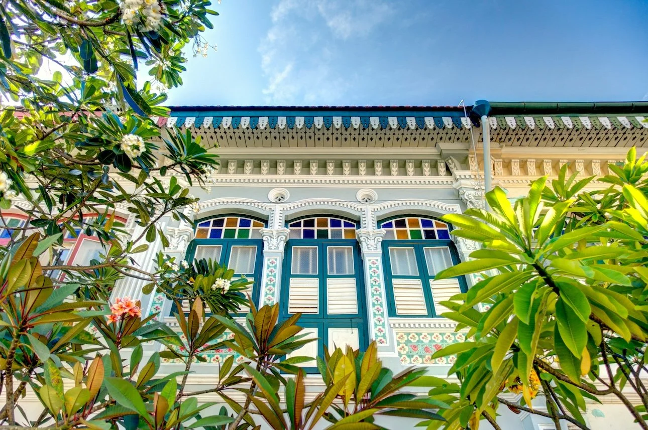 Upside-down view of a colonial-style house with colorful stained glass windows, intricate wooden trims, and surrounded by green foliage against a bright blue sky.