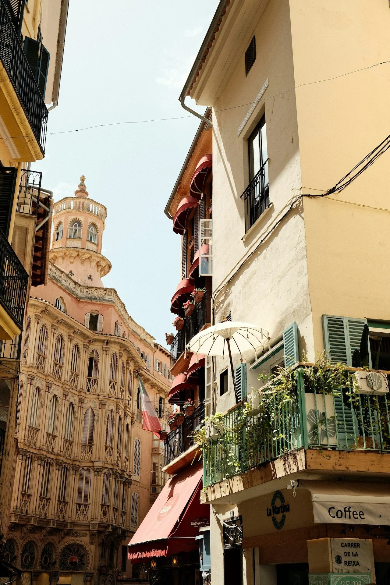Narrow street view in Europe, showing pastel-colored buildings with balconies and awnings, a historic church tower in the background, and a cafe on the corner.