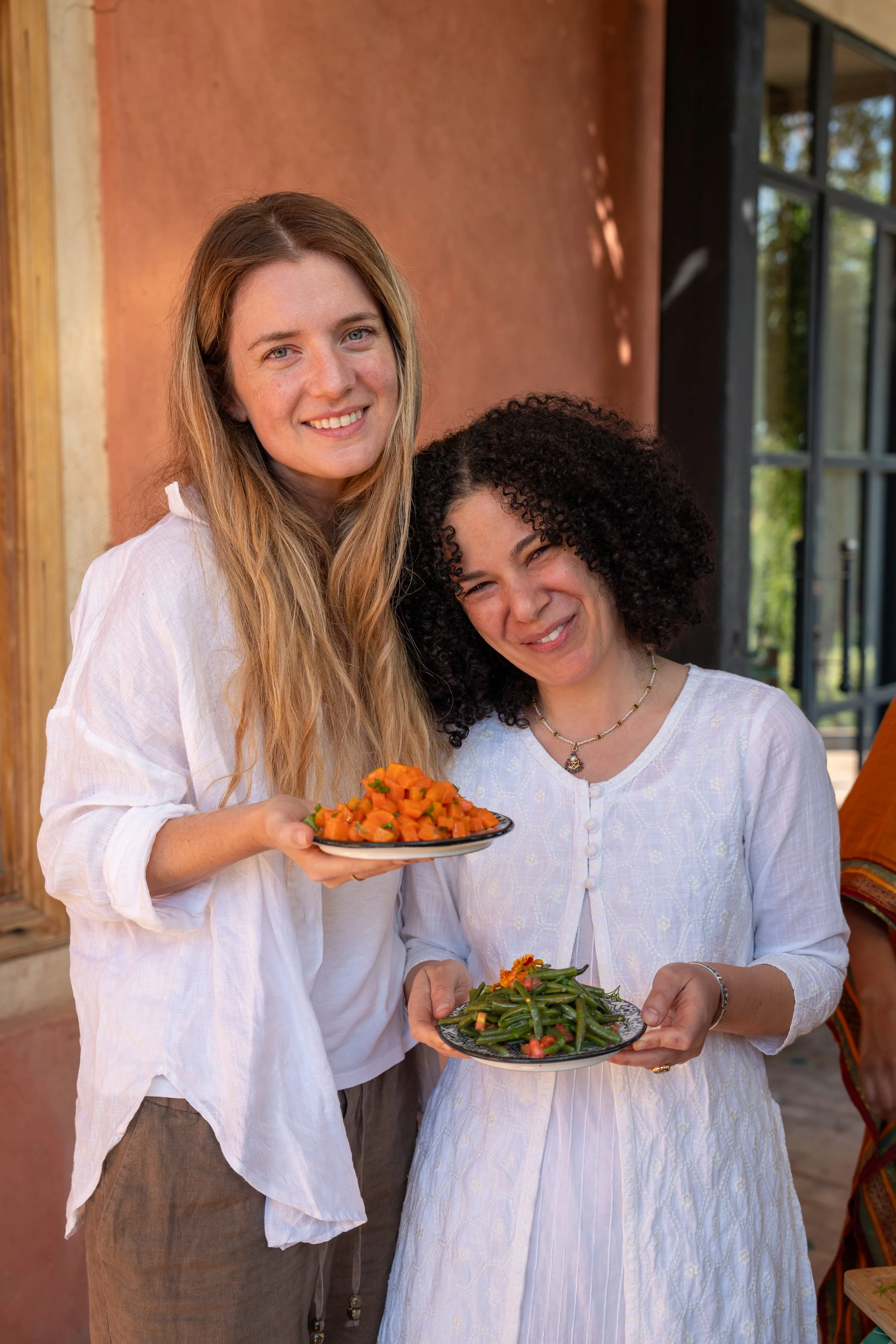 Two women in white clothing holding plates of vegetarian food, including cooked orange vegetables and green beans with flowers on top, standing outdoors.