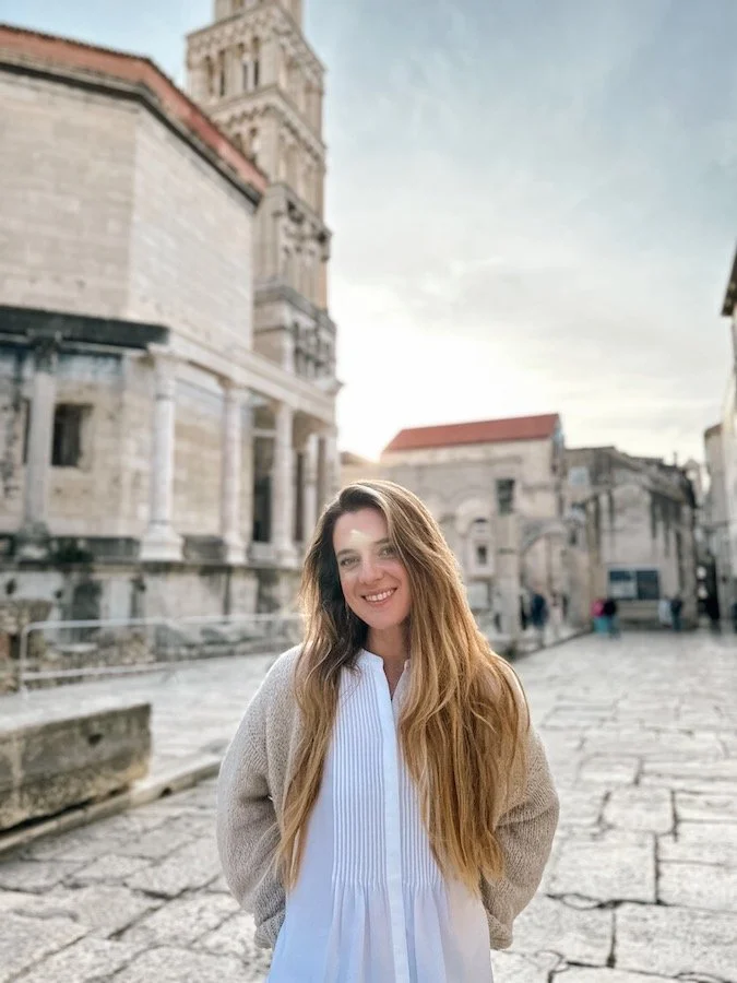 A woman with long blonde hair smiling in front of historic stone buildings on a cobblestone street during daytime.