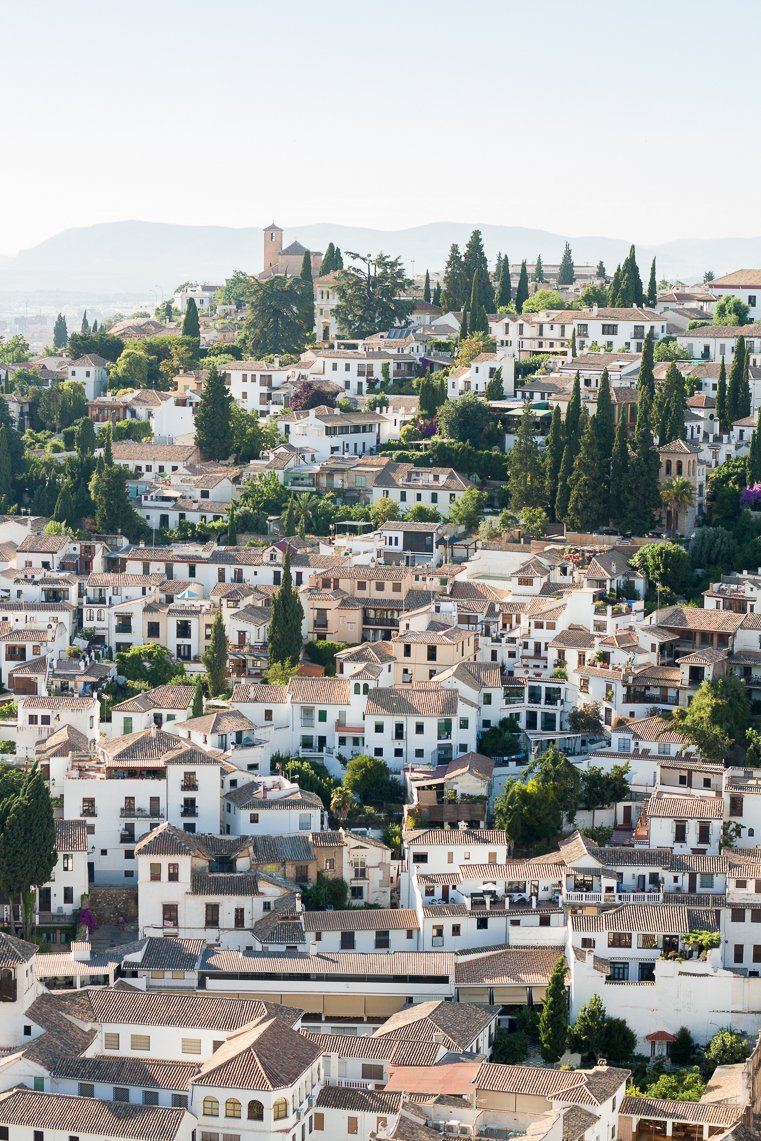 Scenic view of a hillside town with white buildings and red-tiled roofs, surrounded by greenery and cypress trees.