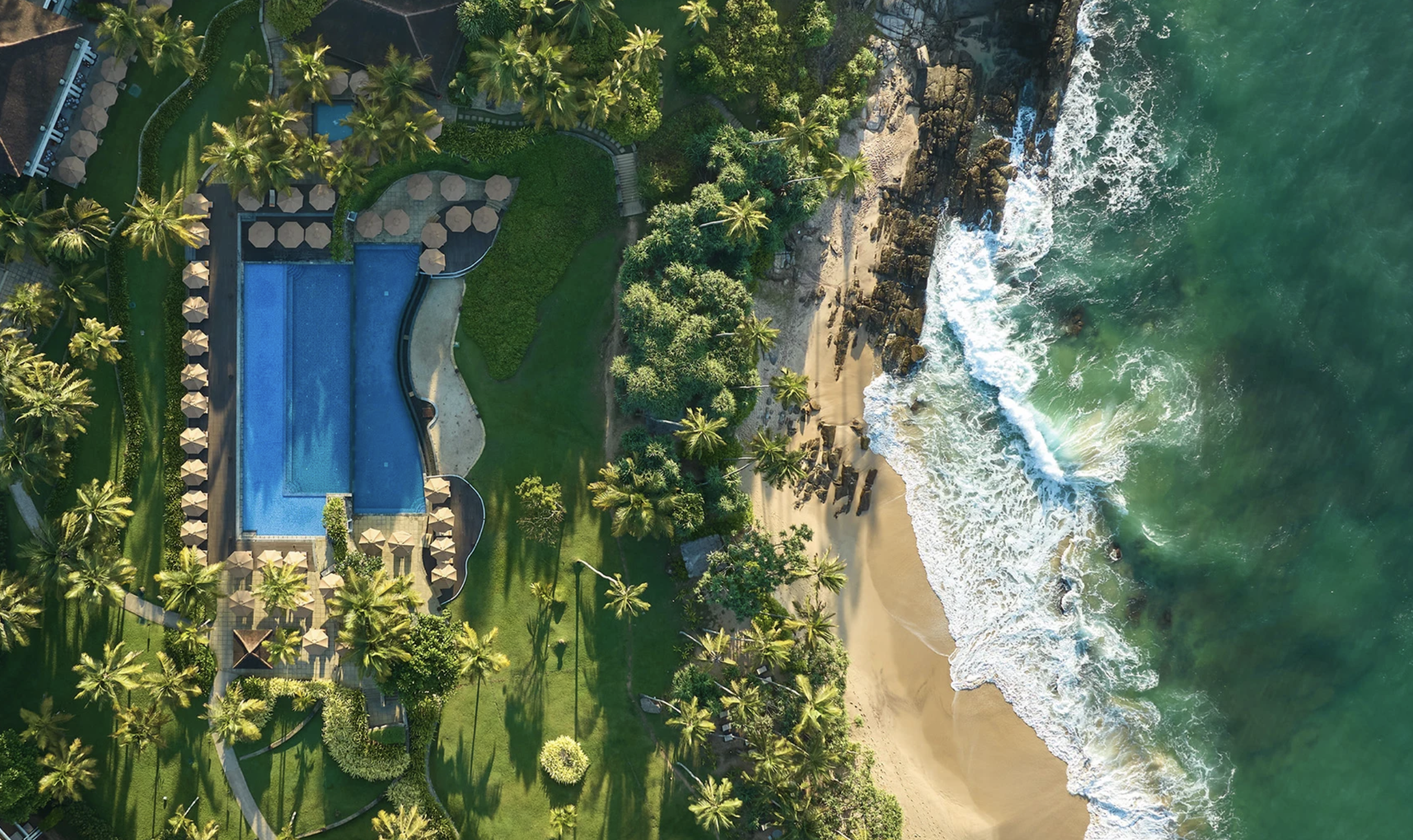 Aerial view of a beachfront resort with a rectangular swimming pool, surrounding umbrellas, palm trees, and adjacent beach with ocean waves.