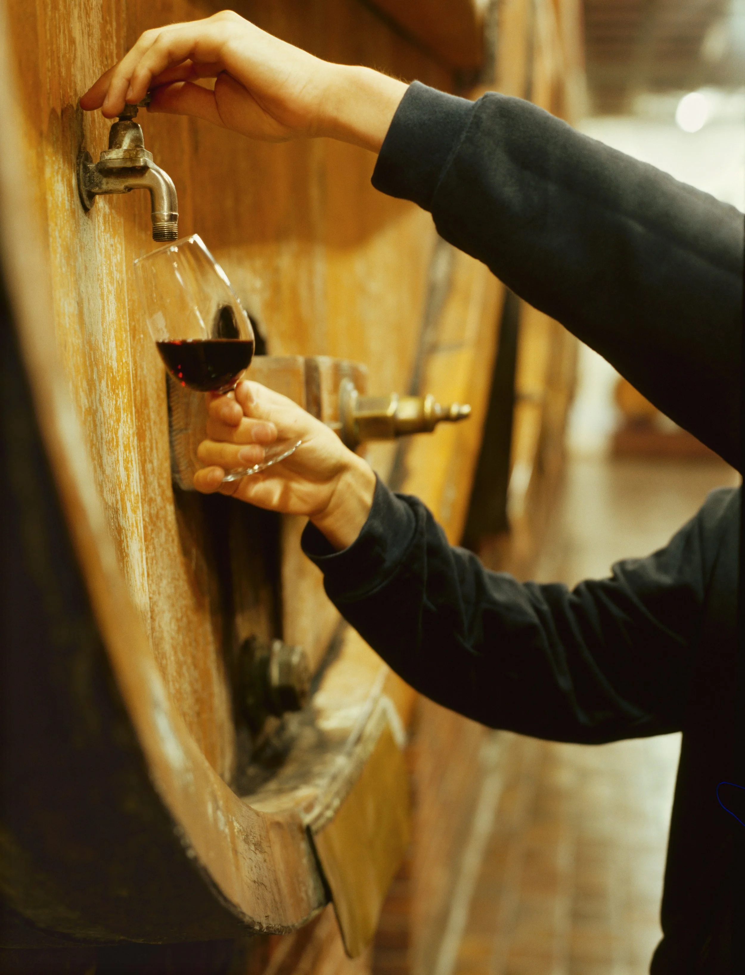 Person filling glass with wine from barrel tap in winery cellar.