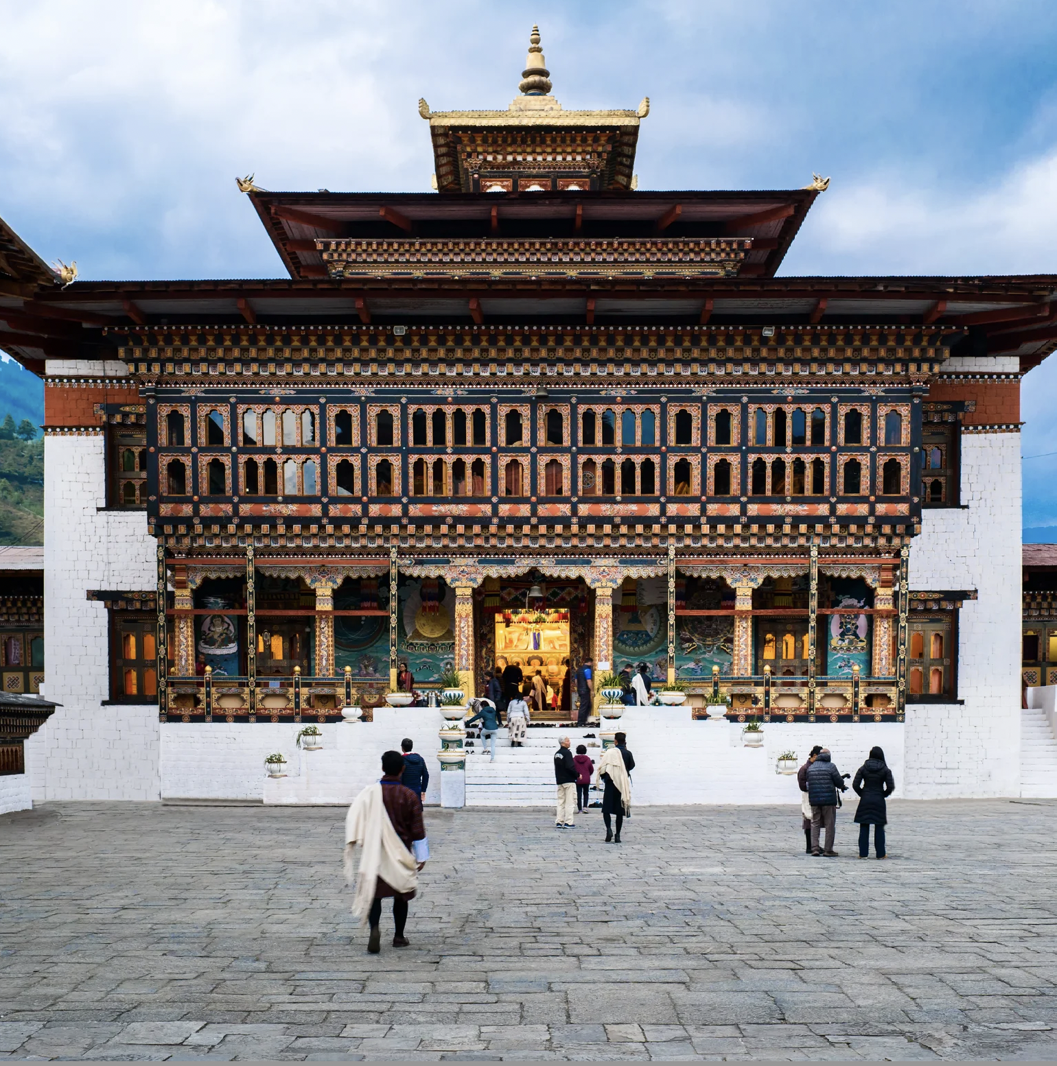 A traditional Bhutanese temple with ornate wooden architecture and a golden spire on top, with people entering and standing outside on a stone courtyard.