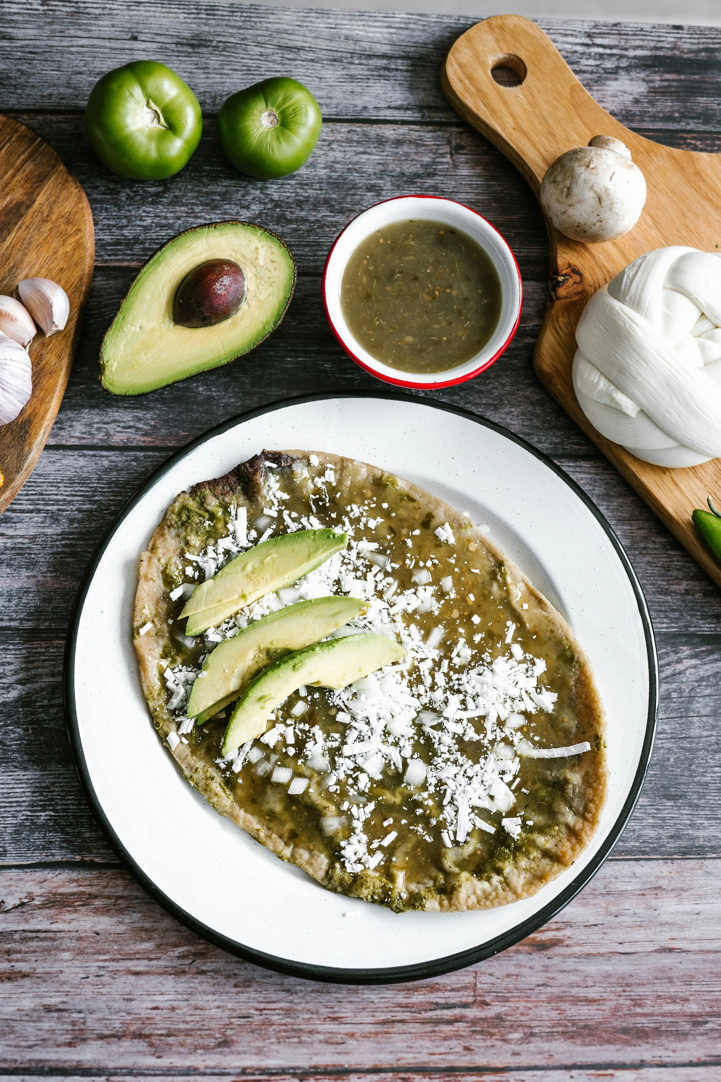 Mexican tlayuda with avocado slices, cheese, green sauce; surrounded by ingredients like avocado, tomatillos, garlic, cheese, mushroom.
