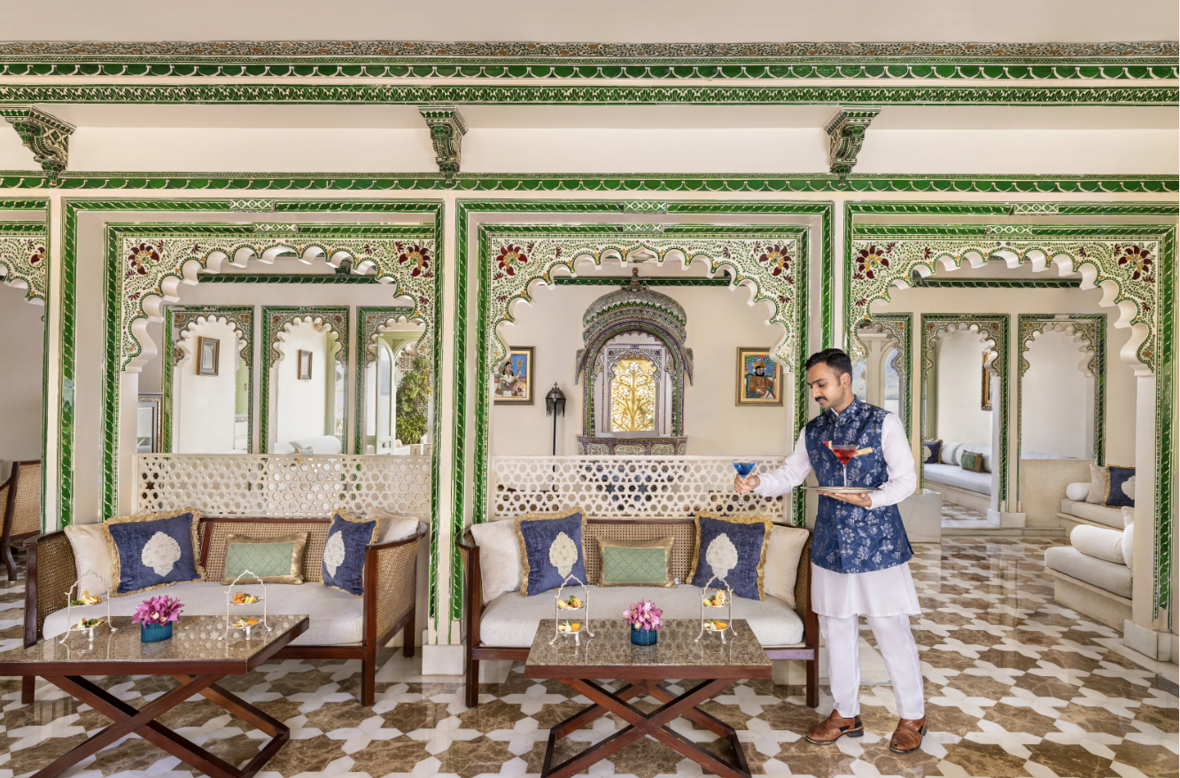 A traditional Indian room with ornate green and white decor, featuring arched doorways and detailed latticework. A server in traditional attire carries drinks on a tray. Elegant seating with colorful cushions is arranged around tables with small floral centerpieces.