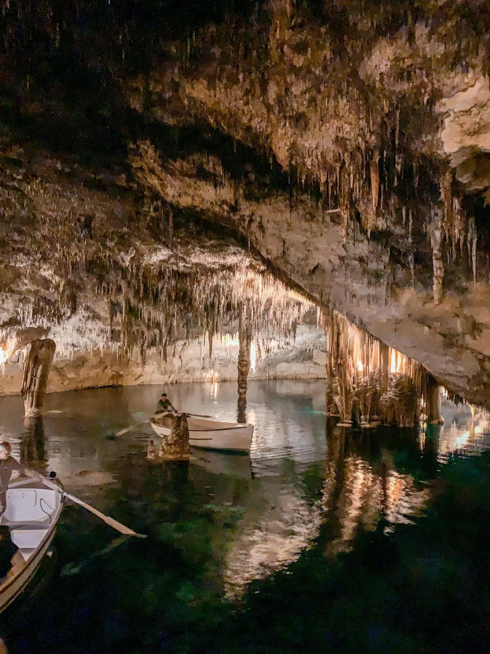 Inside a large cave with stalactites hanging from the ceiling, there are boats on calm water, with people exploring the cave.