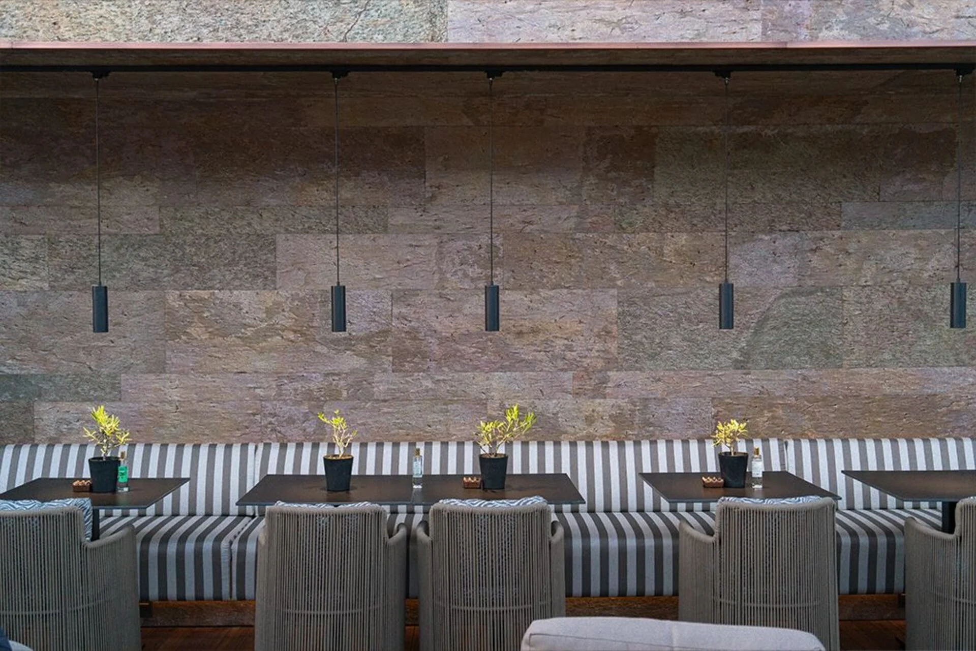 Interior restaurant with a long striped cushioned bench, several small tables, potted plants, water bottles, and hanging pendant lights against a textured stone wall.