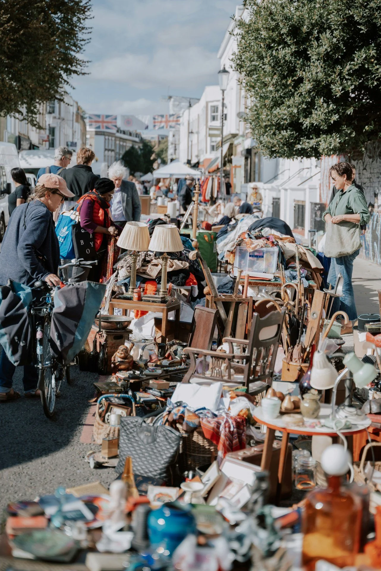 Outdoor street market with various items for sale, including furniture, lamps, and decorative objects, with people browsing and shopping on a sunny day.
