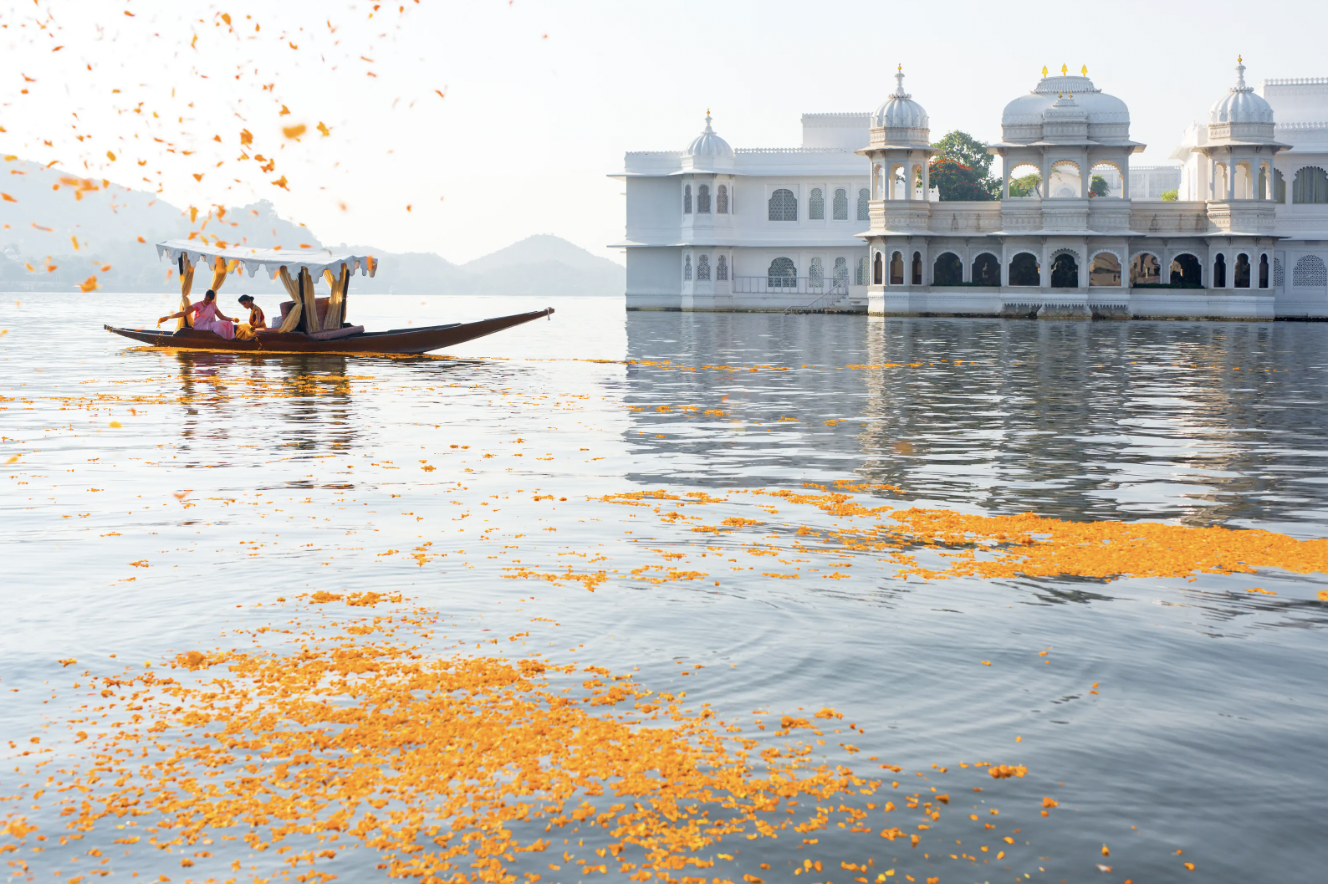 A scenic view of a boat with people on a lake, surrounded by orange flower petals floating on the water, with a white palace in the background.