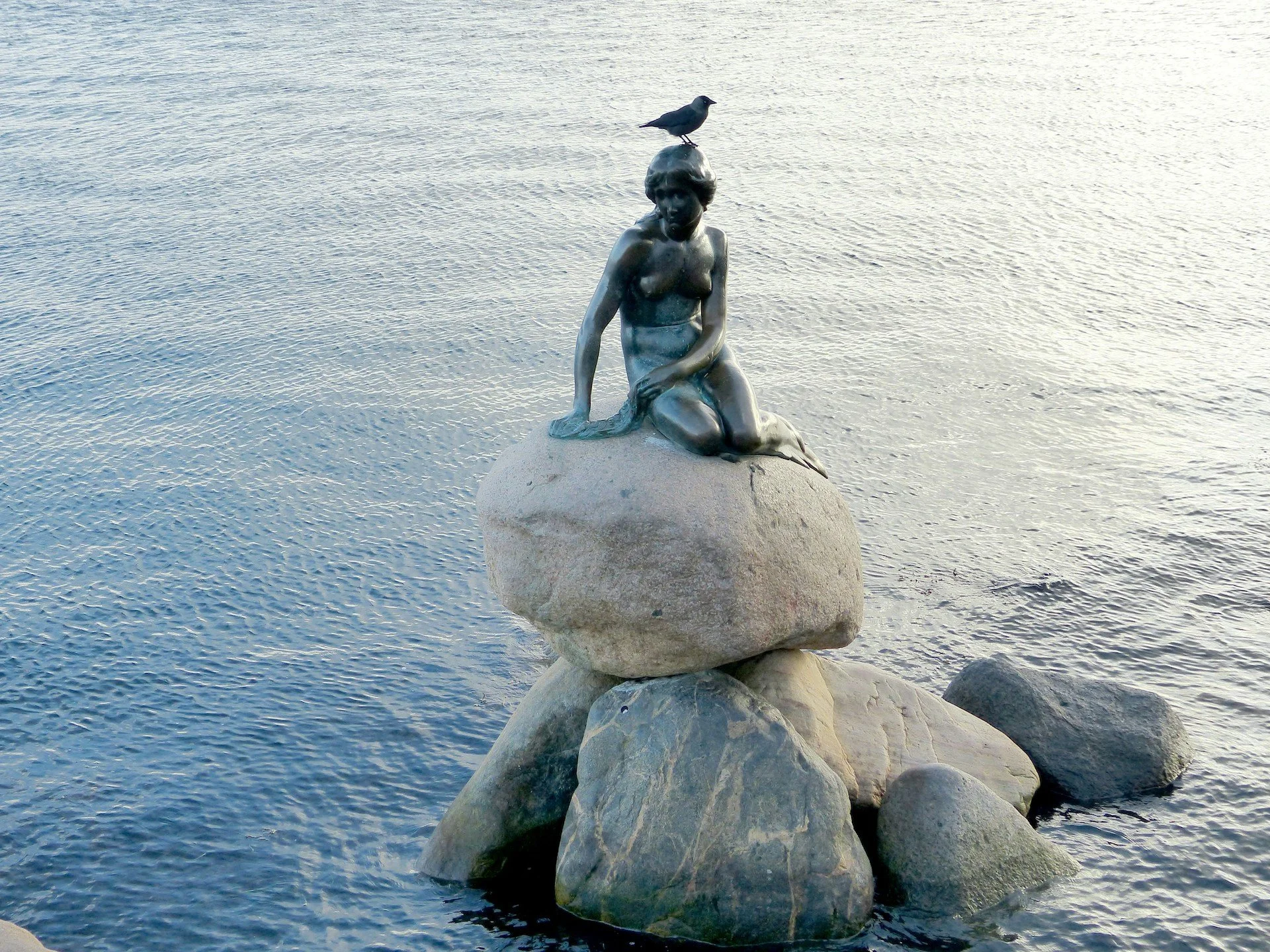 A sculpture of a kneeling woman on a rock in the water, with a bird perched on her head.